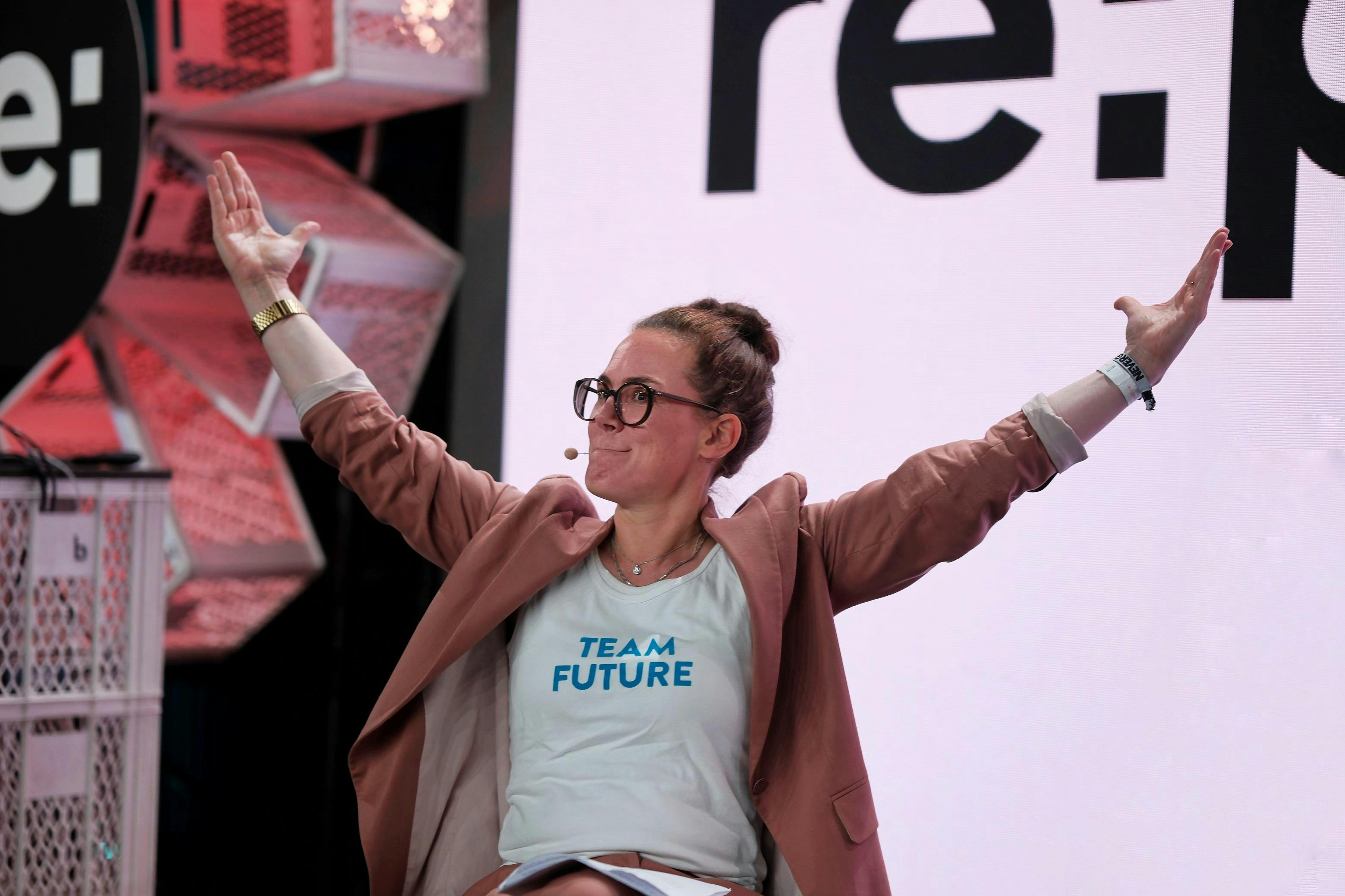 Eine Person mit lachsfarbenem Blazer und weißem Tshirt mit blauer Aufschrift "Team Future" hebt auf einer Bühne sitzend beide Arme.