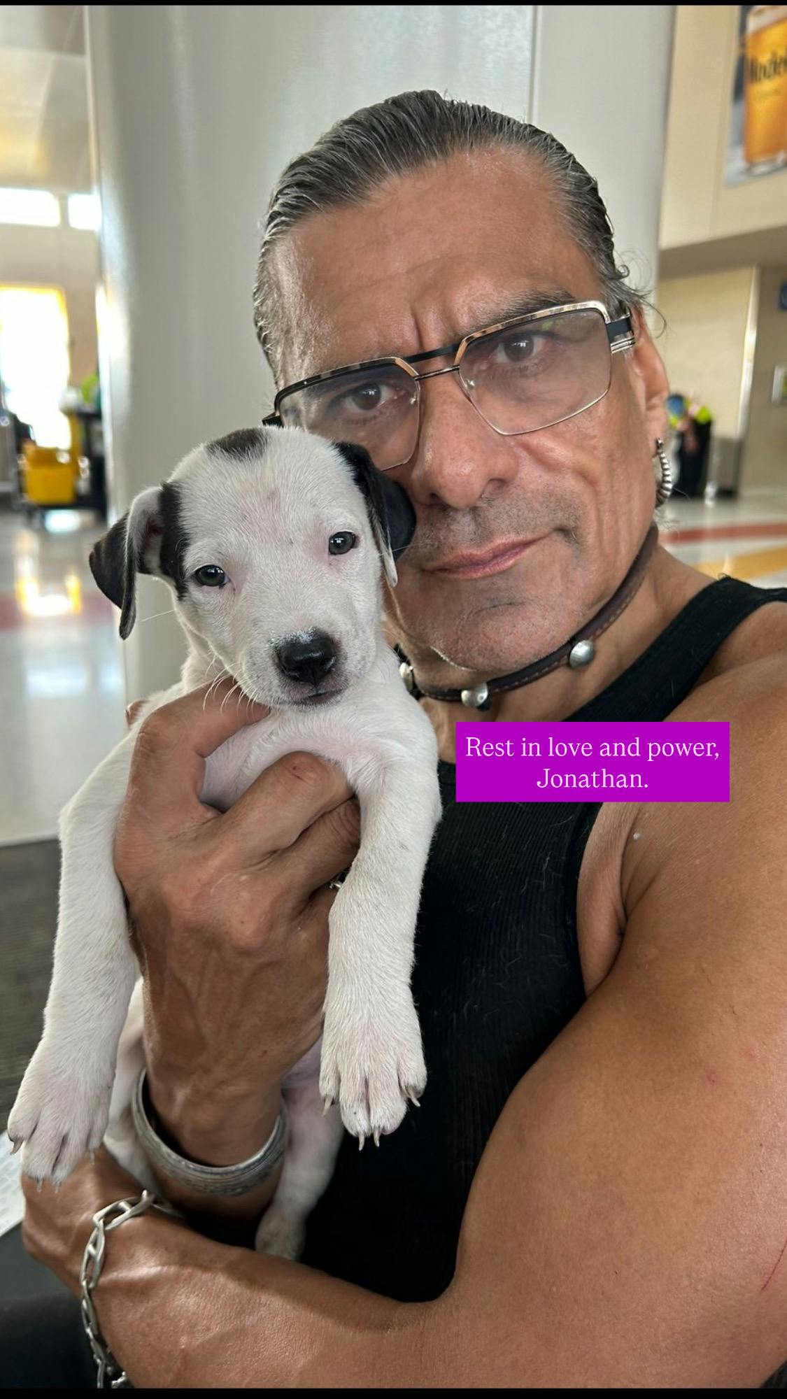 portrait von jonahtan joss mit einem hundewelpen. er trägt eine stärkebrille und schaut direkt in die kamera. im arm hält er ein sehr kleines hundebaby. das hundebaby ist schwarz-weiß und schaut ebenso direkt in die kamera. die unterschrift sagt: rest in love and power, jonathan. ruhe in liebe und power, jonathan.