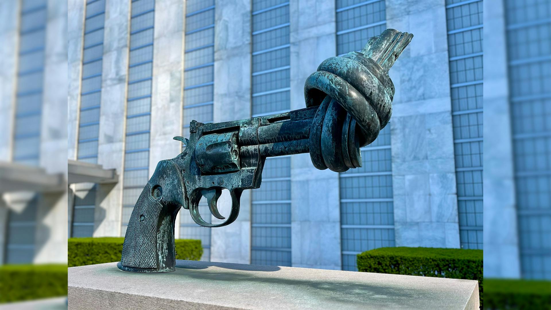 Die grünlich blaue Bronzeskulptur eines Colts, dessen Lauf mit sich selbst verknotet ist. Die Skulptur steht auf einem grauen Sockel. Im Hintergrund links und rechts grüne Hecken. Dahinter ein Gebäude.