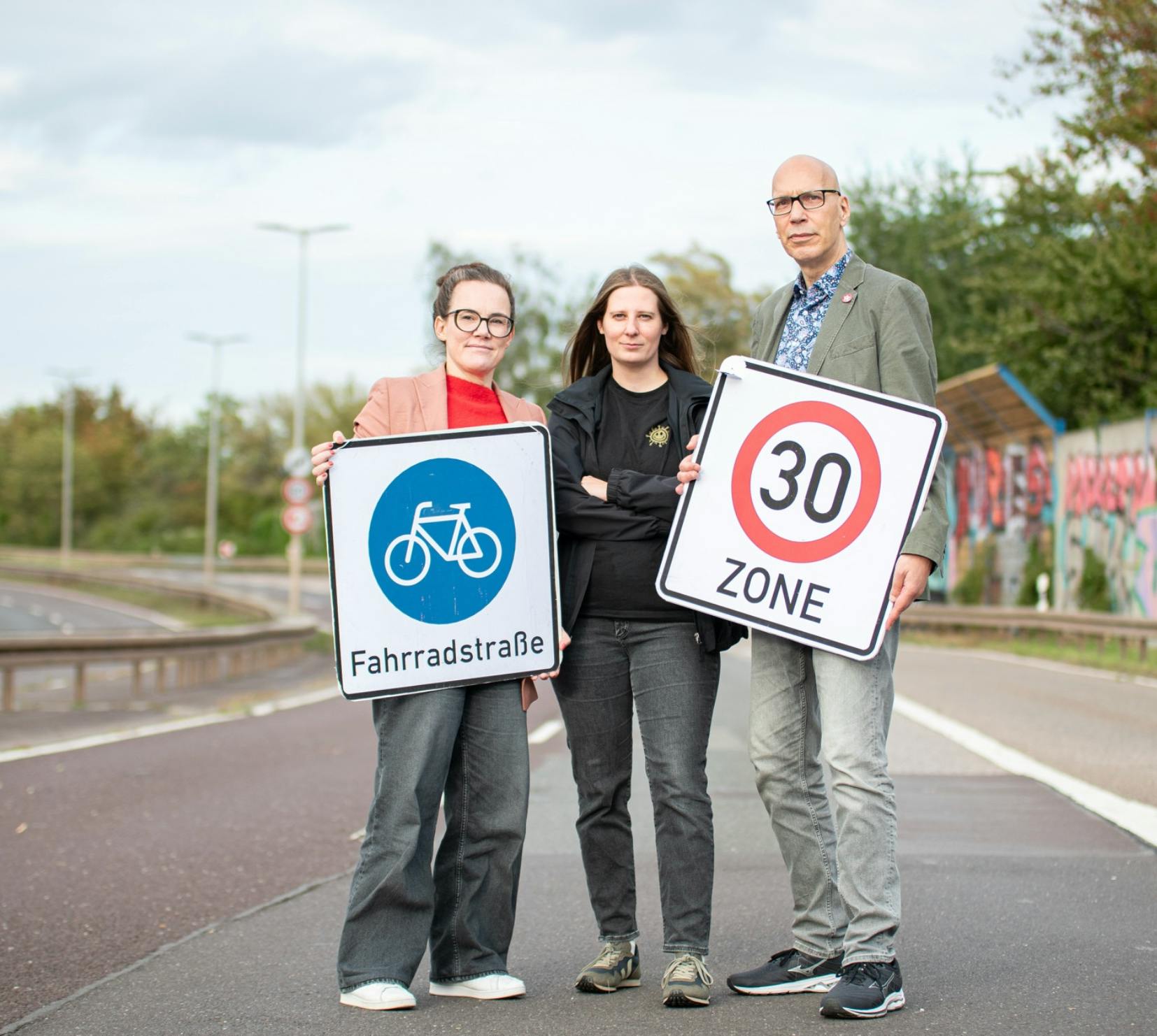 Drei Menschen stehen auf der gesperrten Autobahn. Eine Frau hält ein Schild Fahrradstraße, eine Frau steht mit gekreuzten Armen in der Mitte, der Mann steht rechts und hält das Schild Tempo 30.