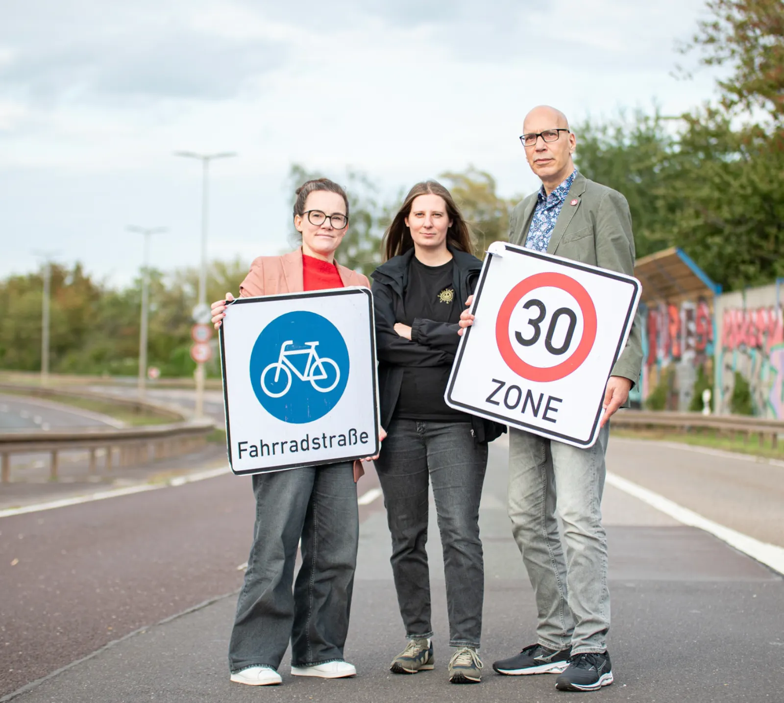 Drei Menschen stehen auf der gesperrten Autobahn. Eine Frau hält ein Schild Fahrradstraße, eine Frau steht mit gekreuzten Armen in der Mitte, der Mann steht rechts und hält das Schild Tempo 30.