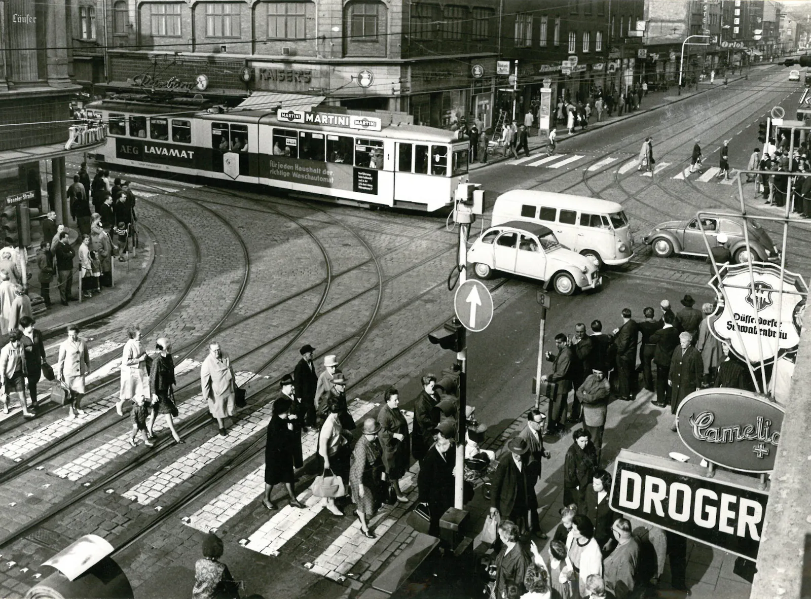 Schwarz-weiß Foto einer städtischen Kreuzung auf der viel los ist. Eine Straßenbahn fährt. Ein VW Bulli und Käfer fahren, viele Leute überqueren einen Zebrastreifen, in der Moder der 1960er Jahre.