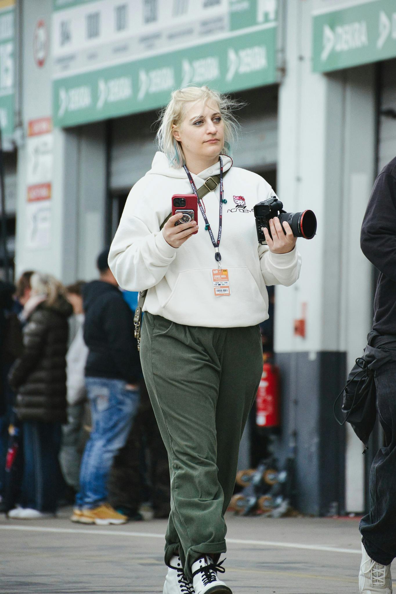 Carconny mit Kamera in der einen Hand, dem Handy in der anderen Hand, läuft im Pitbereich und schaut in die Ferne, hat einen hellen F1-Academy Pullover an und eine grüne Hose, der Hintergrund ist unscharf