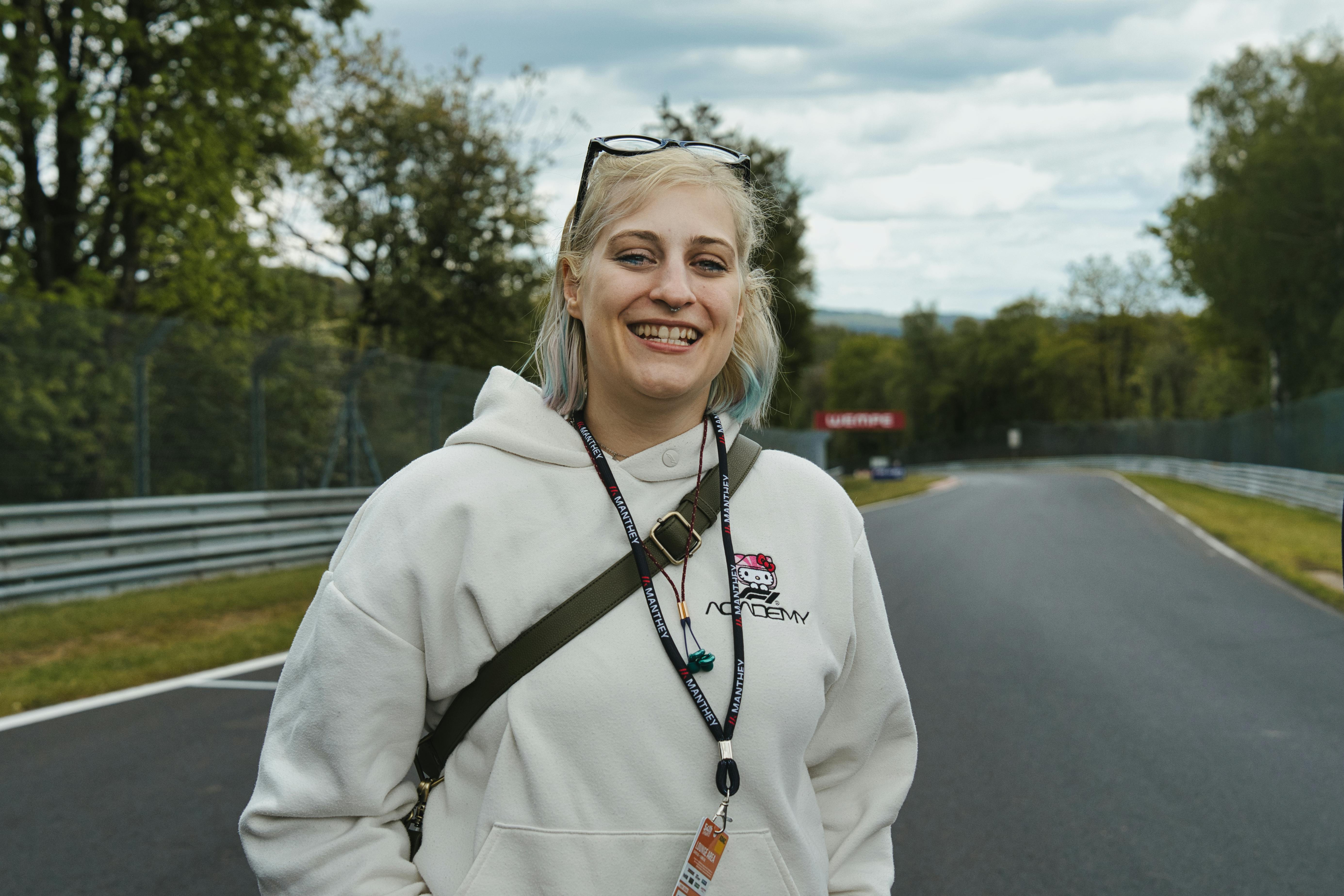 Carconny auf der Rennstrecke des Nürburgrings, eine Blonde Frau mit einem hellen F1-Academy Pullover, lächelnd