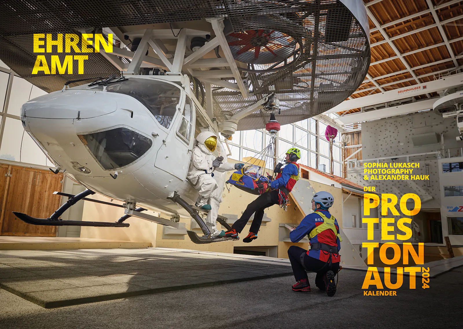 Das Titelblatt des Protestonaut-Kalenders 2024 zum Thema Ehrenamt zeigt Mitglieder der Bergwacht Bad Tölz und den Protestonaut bei einer Hubschrauberübung in der Ausbildungshalle der Bergwacht.