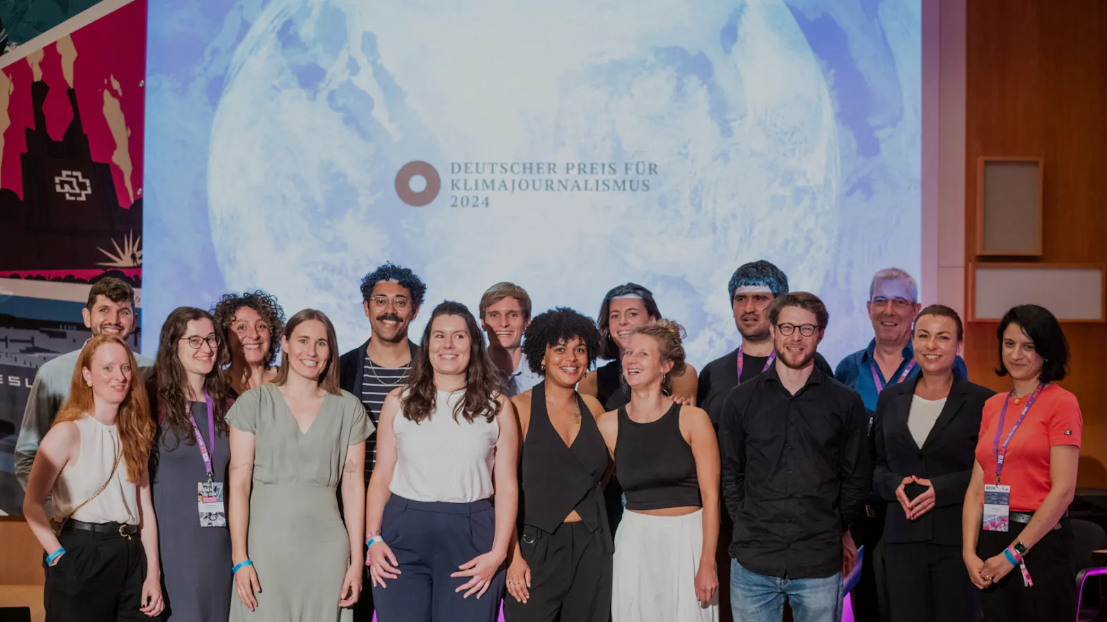 Ein Gruppenbild der Teams, die den Deutschen Preis für Klimajournalismus 2024 gewonnen haben, und Vertreter*innen der Beiträge, die es auf die Shortlist geschafft haben. Die 16 Journalist*innen lachen. Sie sind in zwei Reihen aufgestellt, im Hintergrund sieht man den Schriftzug "Deutscher Preis für Klimajournalismus 2024", darunter stilisiert eine weiße Weltkugel auf blauem Grund.