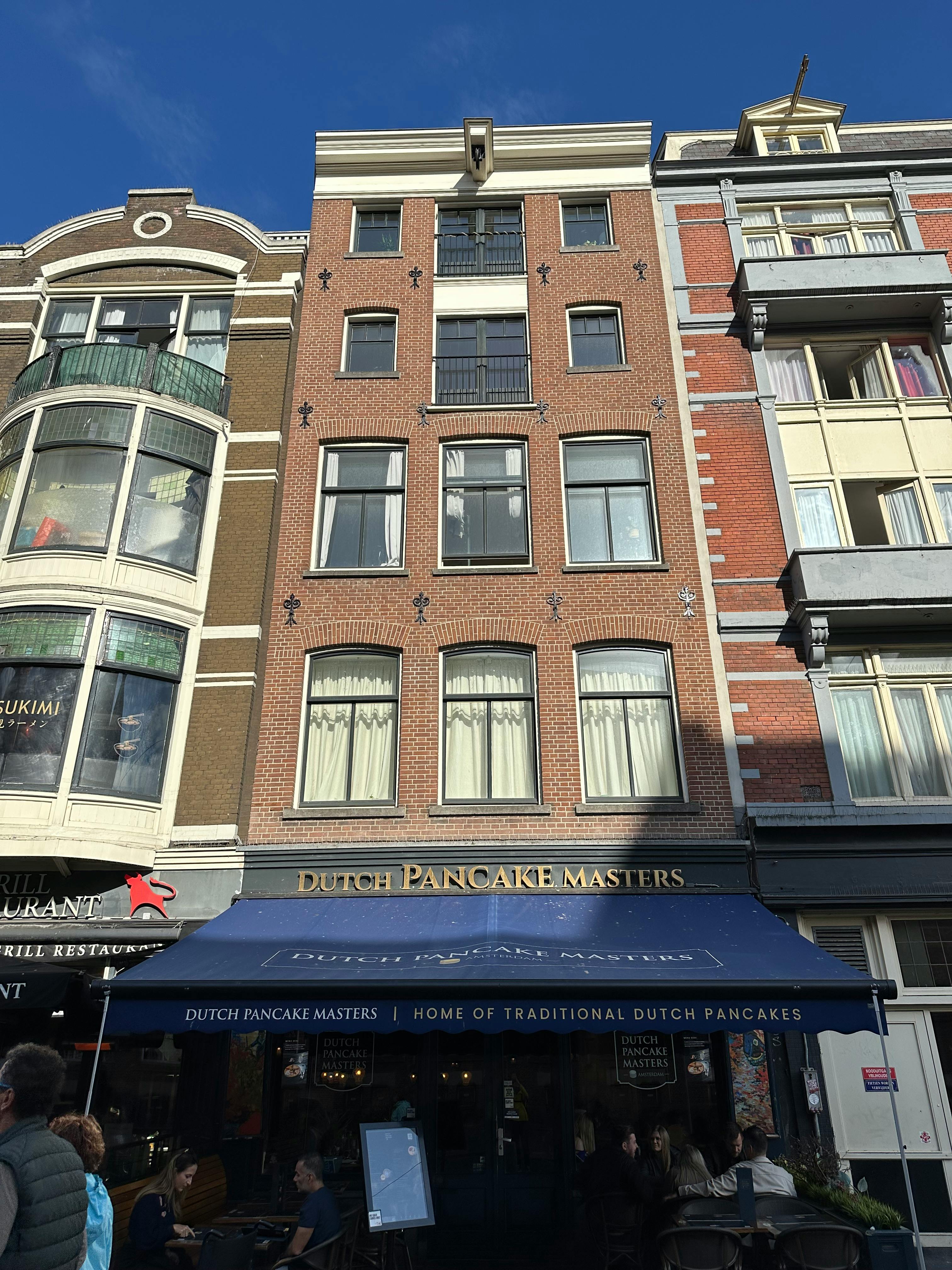 Ein schmales Stadthaus aus roten Backsteinen in der Damrak 44 in Amsterdam. Von der Straße aus sind 4 Etagen zu sehen mit je drei hohen, oben gewölbten Fenstern nebeneinander. Am Dachgiebel ist ein kleiner Ausleger für einen Kran zu sehen. Im Erdgeschoss ist ein Lokal. Das Dutch Pancake Masters. Zur Straße hin ist eine dunkelblaue Markise ausgefahren.