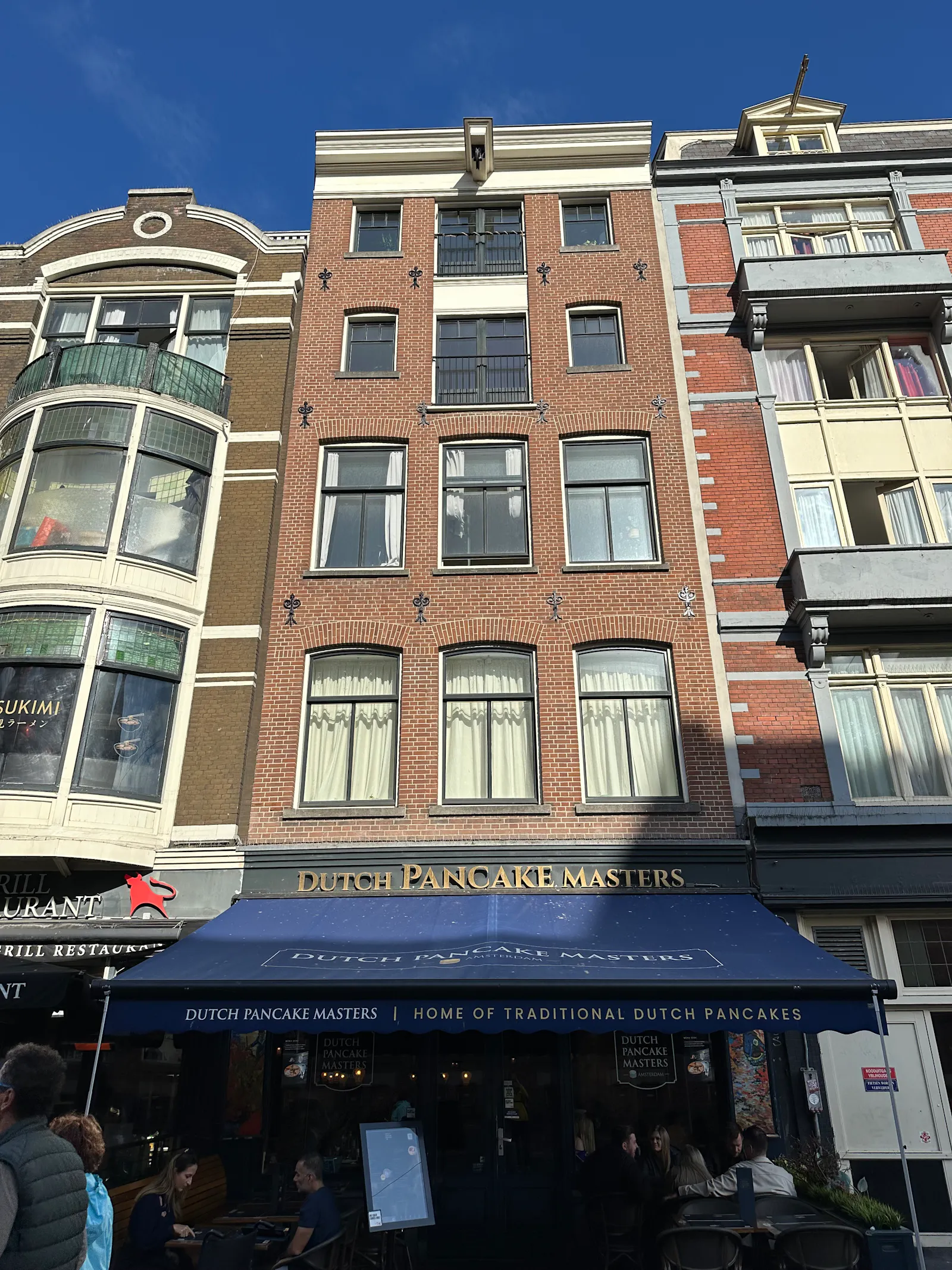 Ein schmales Stadthaus aus roten Backsteinen in der Damrak 44 in Amsterdam. Von der Straße aus sind 4 Etagen zu sehen mit je drei hohen, oben gewölbten Fenstern nebeneinander. Am Dachgiebel ist ein kleiner Ausleger für einen Kran zu sehen. Im Erdgeschoss ist ein Lokal. Das Dutch Pancake Masters. Zur Straße hin ist eine dunkelblaue Markise ausgefahren.