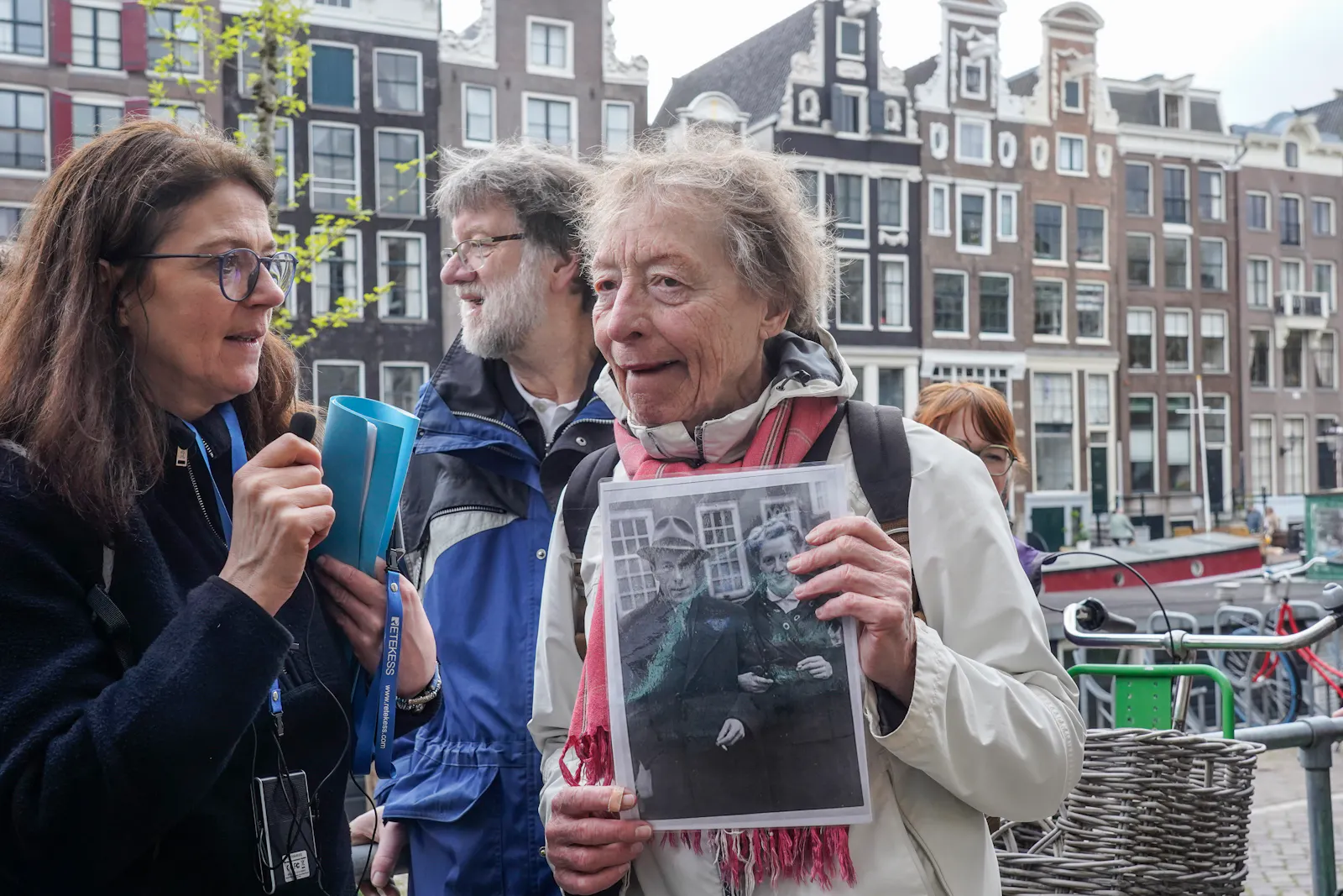 Im Hintergrund sind die typischen niederländischen Grachtenhäuser zu sehen. Im Vordergrund eine über 80-jährige Niederländerin, die ein schwarzweiß Foto ihrer Eltern in der Hand hält. Rechts von ihr eine jüngere Frau mit langen braunen Haaren, die ein kleines Mikrofon in der Hand hält.