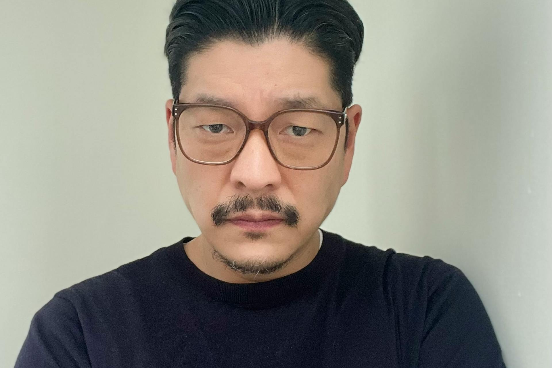 Bill Yom trägt einen schwarzen Sweater vor hellem Hintergrund und schaut ernst in die Kamera.