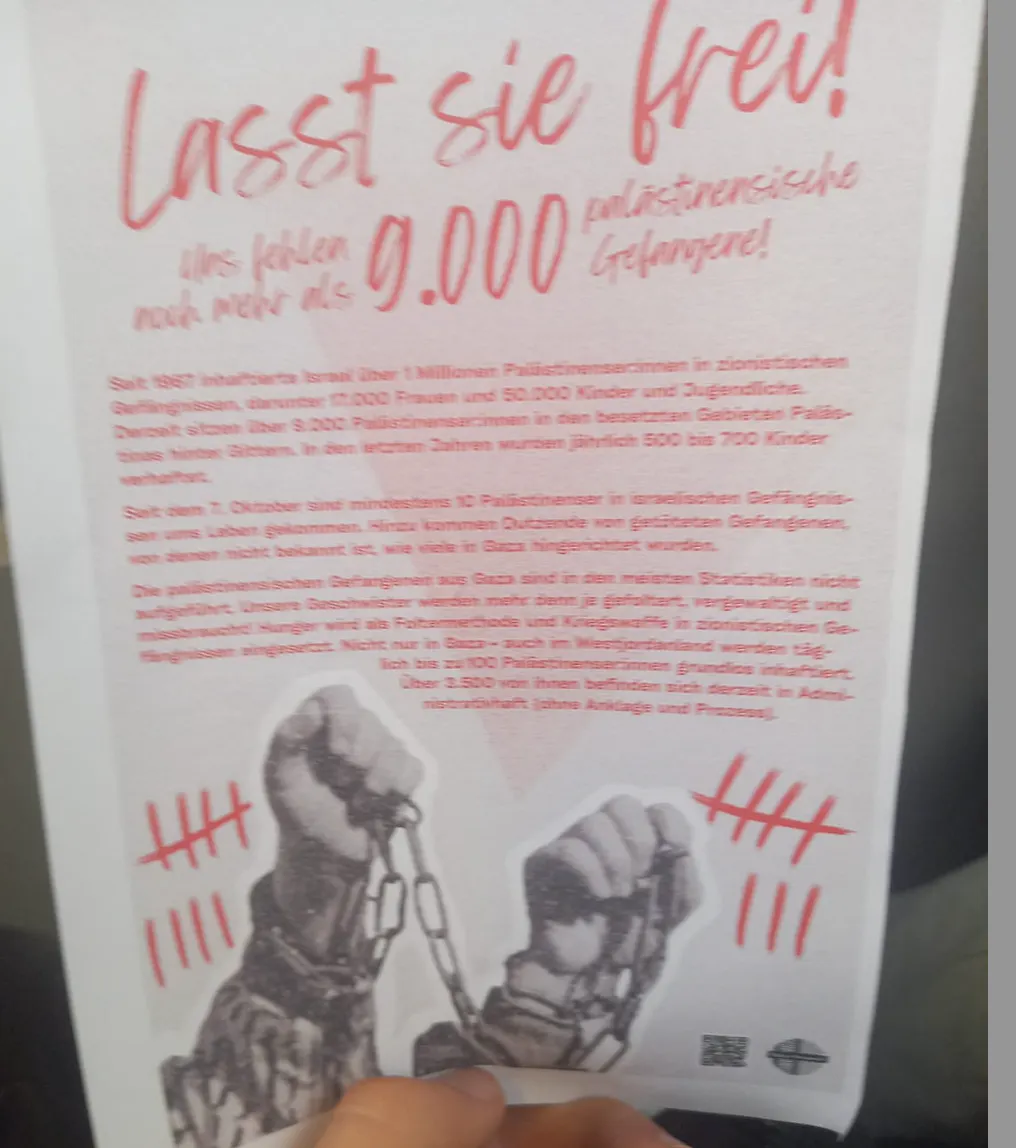 Ein Flyer mit der Überschrift in roter Handschrift: „Lasst sie frei! Uns fehlen noch mehr als 9.000 palästinensische Gefangene!“. Darunter folgt ein mehrspaltiger Text in roter Schrift, der Israel beschuldigt, seit 1967 über eine Million Palästinenser:innen, darunter Frauen und Kinder, willkürlich in „zionistischen Gefängnissen“ inhaftiert zu haben. Es wird von Folter, Hunger, Gewalt und systematischer Unterdrückung gesprochen. Eine Zahl von 9.000 aktuellen Gefangenen wird genannt. Im unteren Bereich des Flyers sind zwei schwarz-weiße Hände mit Ketten dargestellt, die in einer Geste des Protests geballt sind. Daneben befinden sich rote Strichlisten (Kerbmarkierungen), wie man sie zur Zählung verwendet. Unten rechts ist ein kleines Logo einer Organisation sowie ein QR-Code zu sehen.