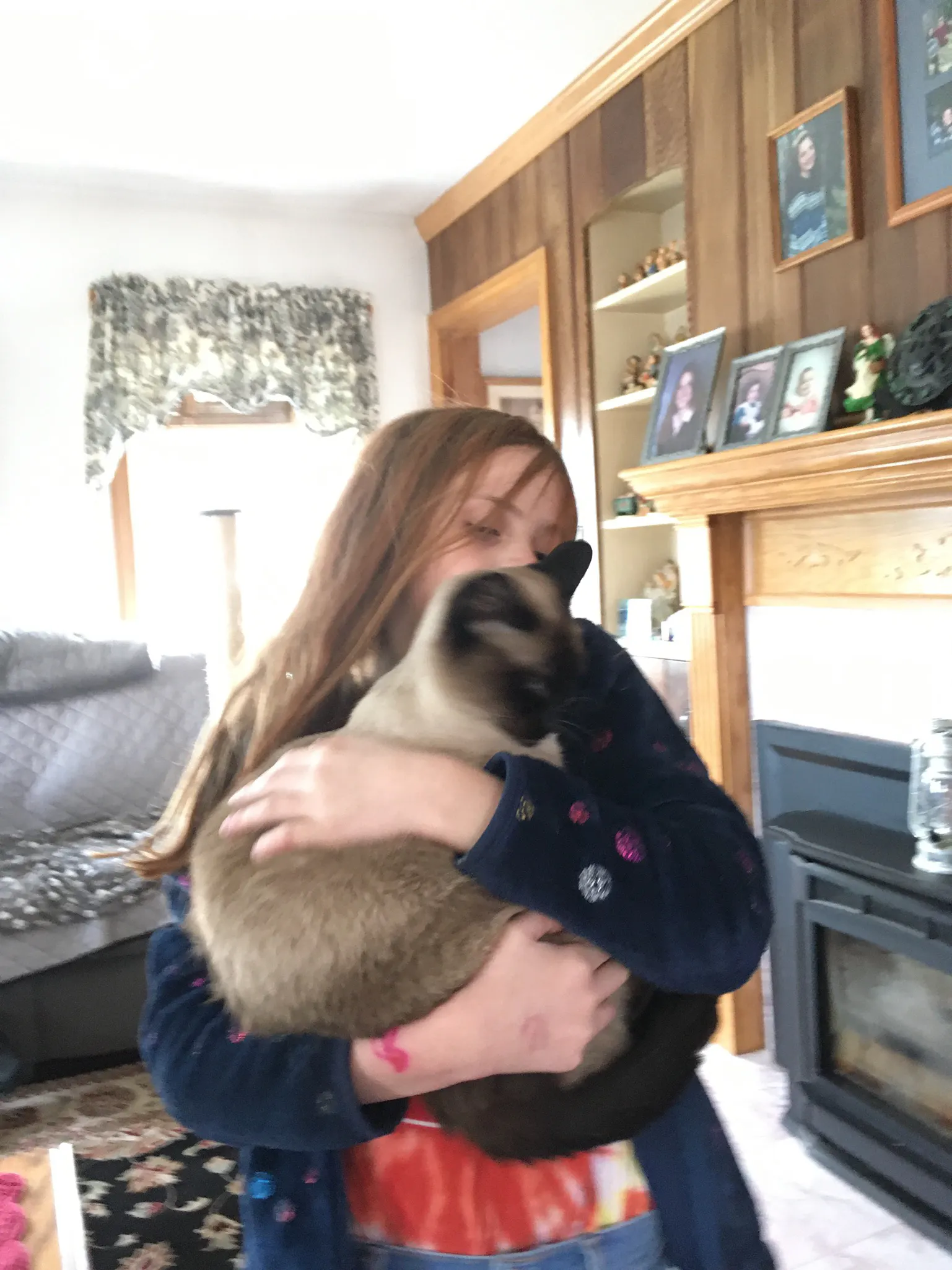A Siamese cat dances with my niece during the early days of the COVID-19 lockdown. The grey love seat is in the background with a black fireplace to the right of niece and Madra in her arms. My niece is blonde with a blue jacket and tie dye Skittles shirt. Pictures of my sister adorn the mantle above the fireplace.