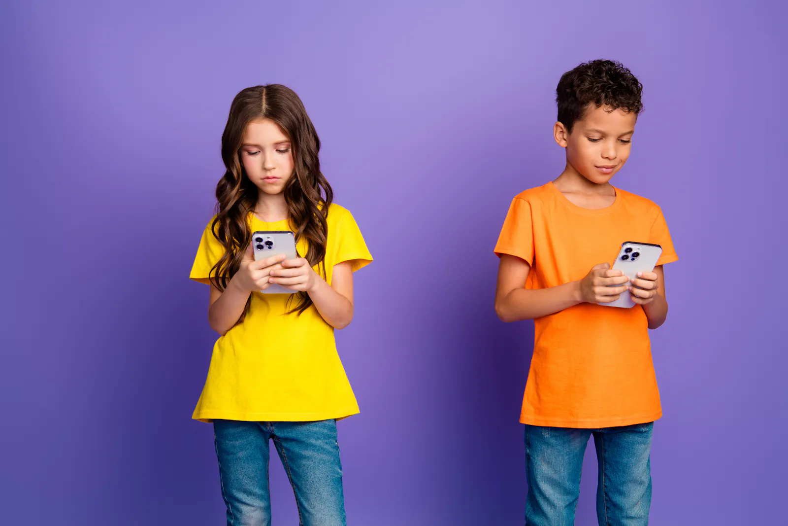Das Bild zeigt zwei Kinder vor einem lila Hintergrund. Links steht ein Mädchen mit langen braunen Haaren in einem gelben T-Shirt. Rechts steht ein Junge mit lockigen Haaren in einem orangefarbenen T-Shirt. Beide schauen konzentriert auf ihre Smartphones.