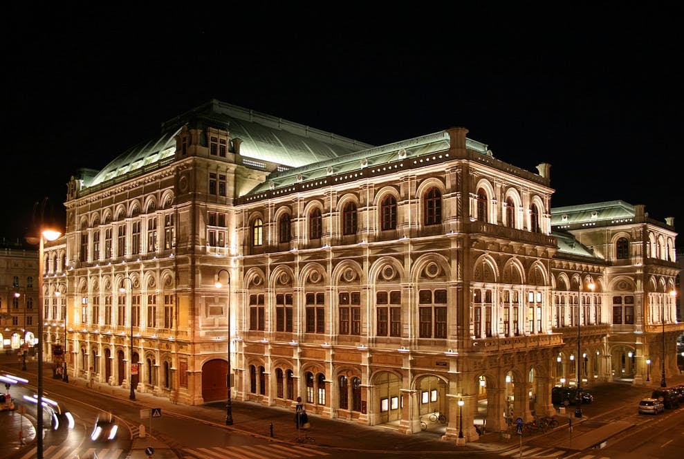Das Bild zeigt die Wiener Staatsoper bei Nacht. Das Gebäude ist eindrucksvoll beleuchtet und präsentiert sich im Stil der Neorenaissance. Autos fahren auf der Straße davor, und die Fassade ist hell angestrahlt.