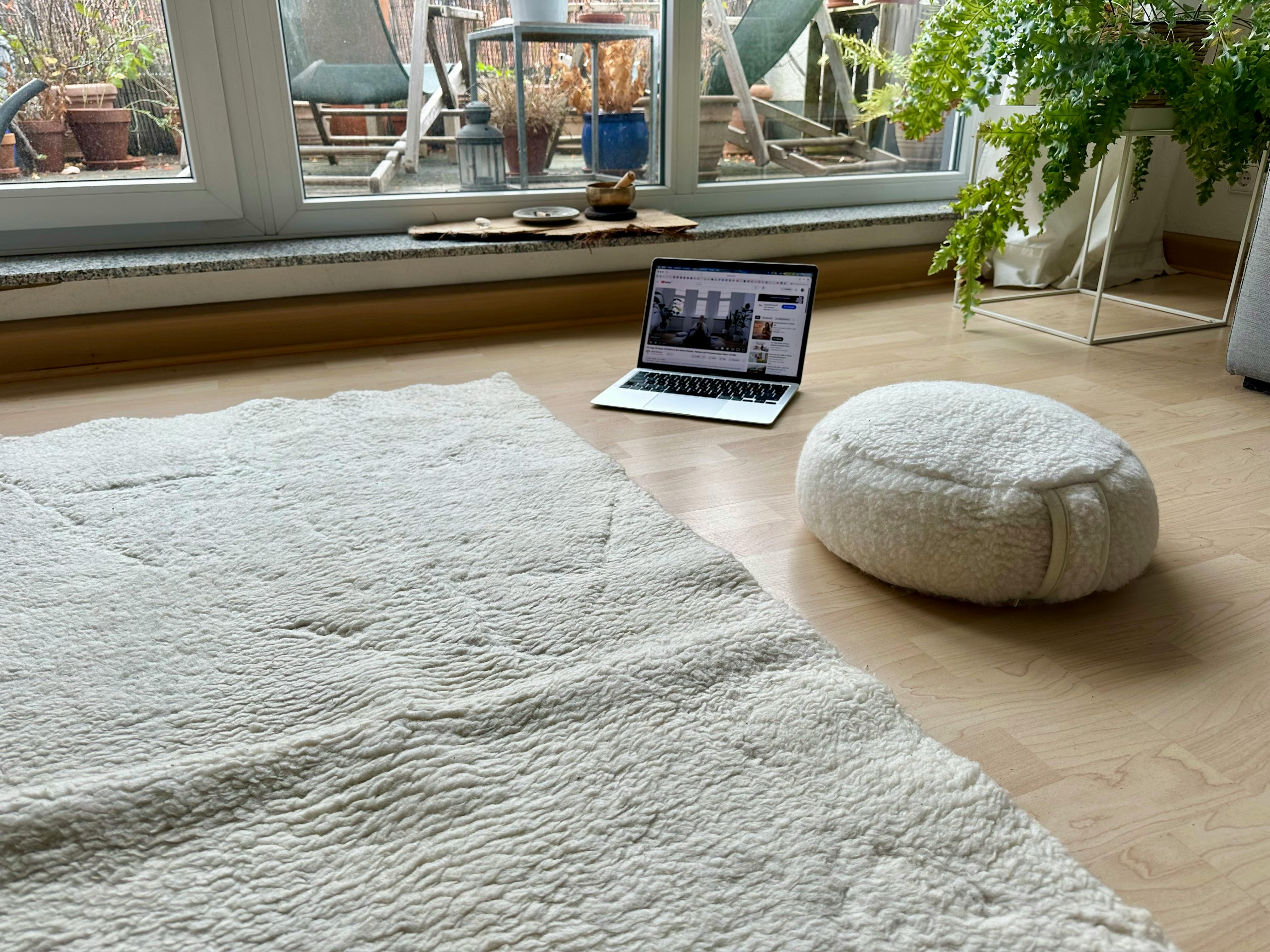 Schurwoll-Yogamatte auf dem Boden. Daneben ein Schurwoll-Meditationskissen sowie ein Laptop, auf dem ein Yogavideo zu sehen ist.