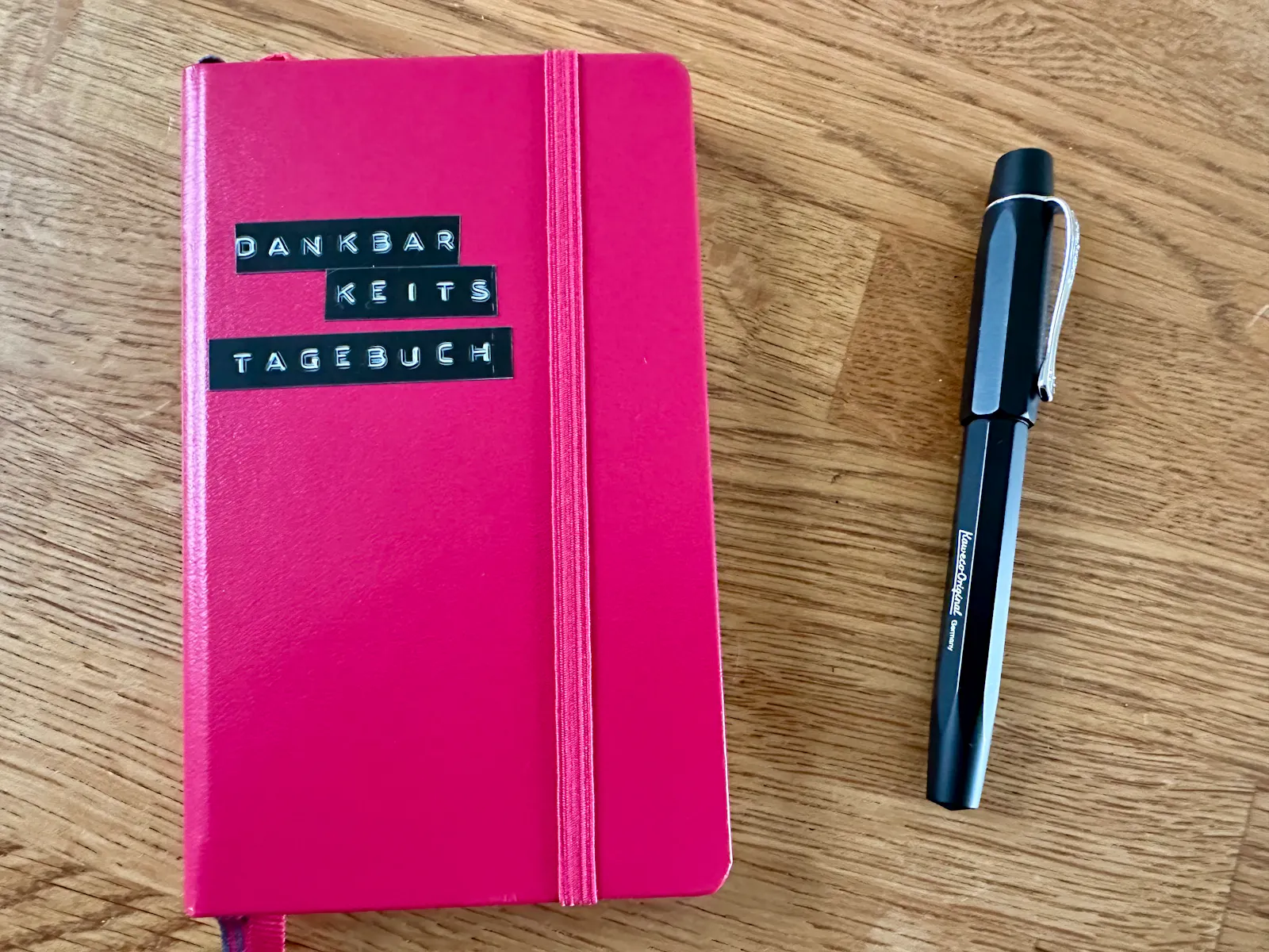 DIN A6 Notizbuch in pink, darauf ein schwarzes Klebeetikett "Dankbarkeitstagebuch". Daneben ein schwarzer Füller.