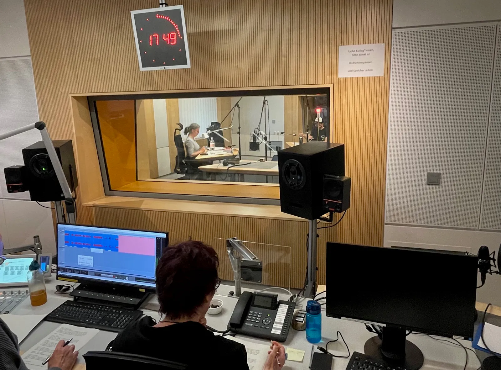 Foto von den Studioaufnahmen, Blick von oben auf Tisch mit Mischpult, Bildschirmen und Boxen, mit Fenster in der Wand und Durchblick zu den Sprecher*innen-Plätzen und Mikrophonen.