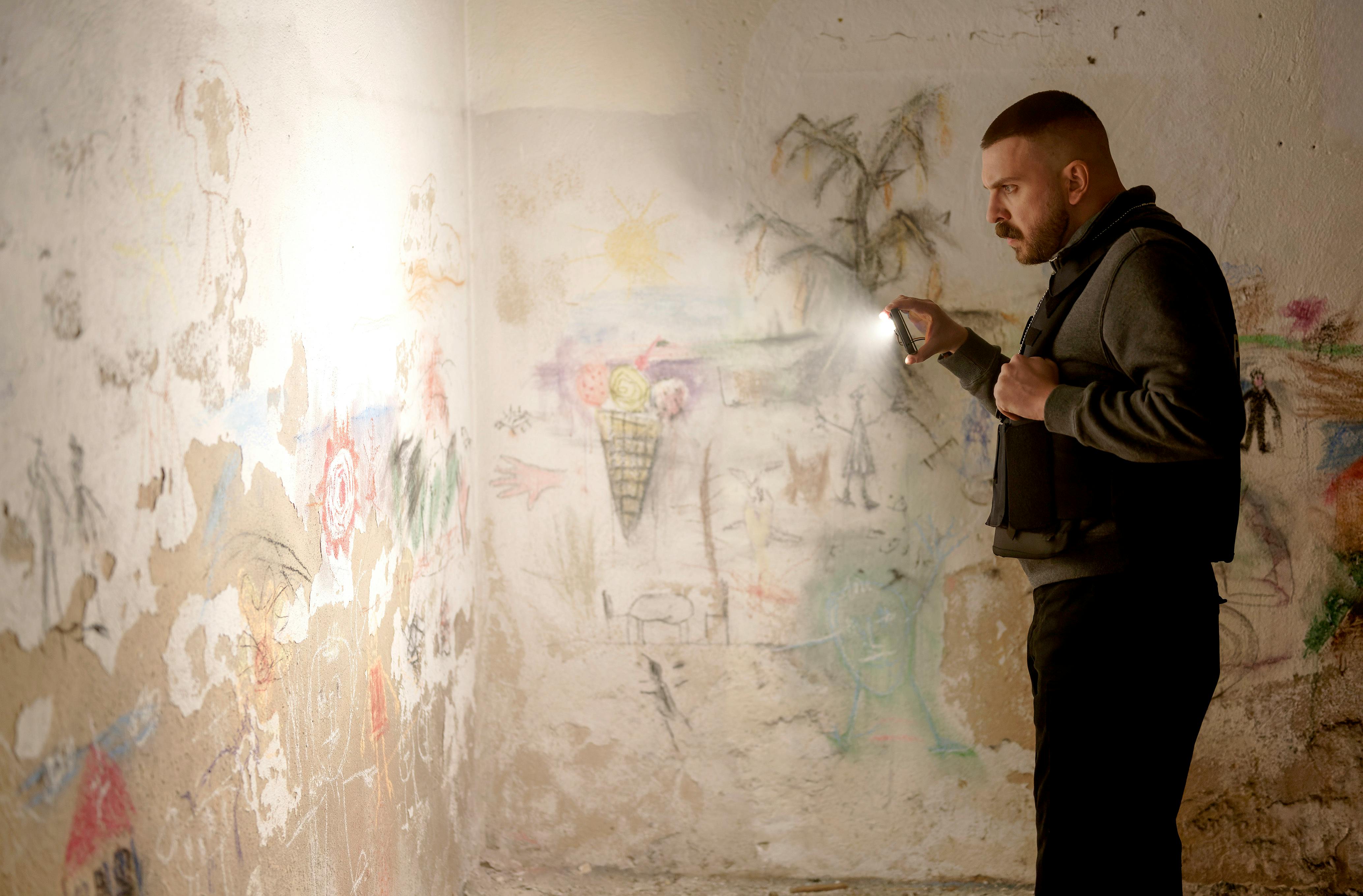 Hamza Kulina (Edin Hasanovic) in einem Nebenraum der Lichtsekte, mit Taschenlampe schaut er sich Wandkritzeleien an © HR/Sommerhaus/Tatiana Vdovenko