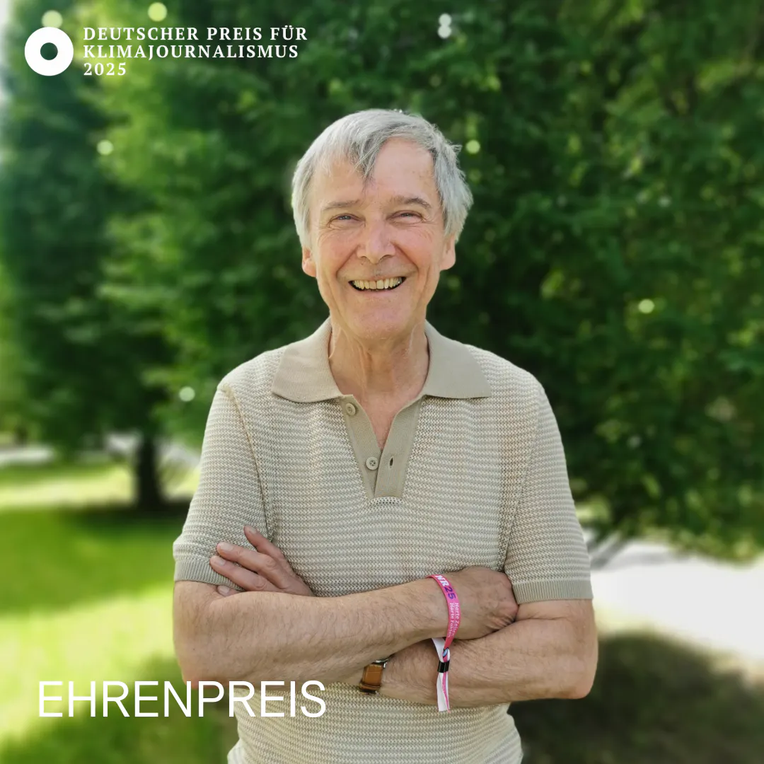 Das Bild zeigt Ehrenpreis-Träger Georg Ehring, er trägt ein beiges Poloshirt, hält die Arme verschränkt und lächelt. Im Hintergrund sind eine Wiese und Bäume zu sehen.