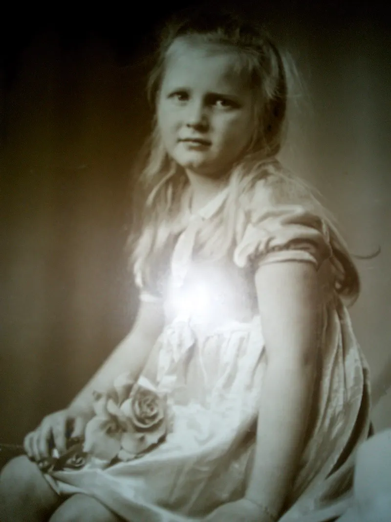 A photo of a girl about eight years old sitting facing to the left with her face turned towards us. She is wearing a puff sleeved dress and has long blond hair. She is holding a rose in her lap. Her expression could be sad, or wondering why she has to sit like this.
