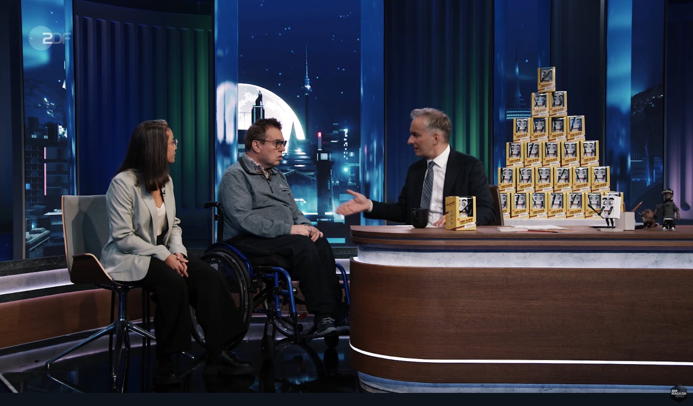 Bühne einer Fernsehshow, hinter einem Tisch sitzt Jan Böhmermann, neben ihm Jürgen Linnemann, daneben Soraia Da Costa Batista. Rechts steht eine Pyramide aus Grundi-Figuren. Im Hintergrund ist ein Nachthimmel mit Mond projeziert.