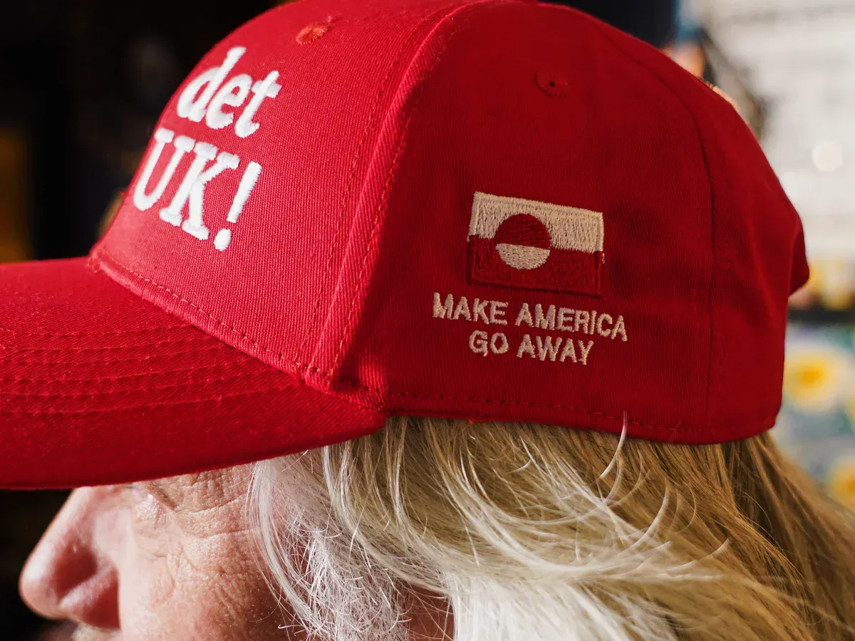 Eine rote cap mit weißer Schrift, auf der vorne “Nu det NUUK!” Und an der Seite unter einer Grönland-Flagge “Make america go away” steht