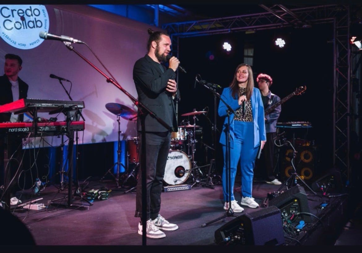 Das Bild zeigt Marco und Jasmin auf der Bühne während ihrer Moderation des Credo-Collab- Abends. Marco trägt einen schwarzen Anzug und hält ein Mikrofon in der Hand, Jasmin trägt einen blauen Hosenanzug und interagiert mit dem Publikum.