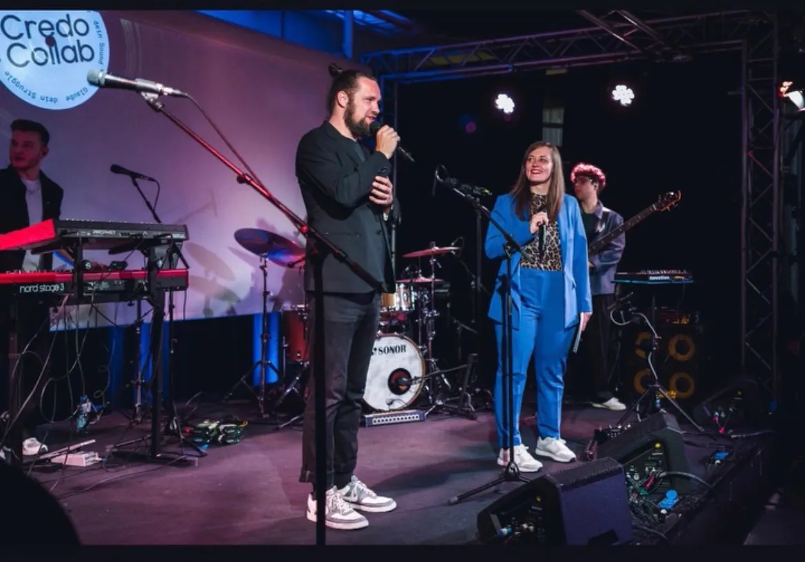 Das Bild zeigt Marco und Jasmin auf der Bühne während ihrer Moderation des Credo-Collab- Abends. Marco trägt einen schwarzen Anzug und hält ein Mikrofon in der Hand, Jasmin trägt einen blauen Hosenanzug und interagiert mit dem Publikum.