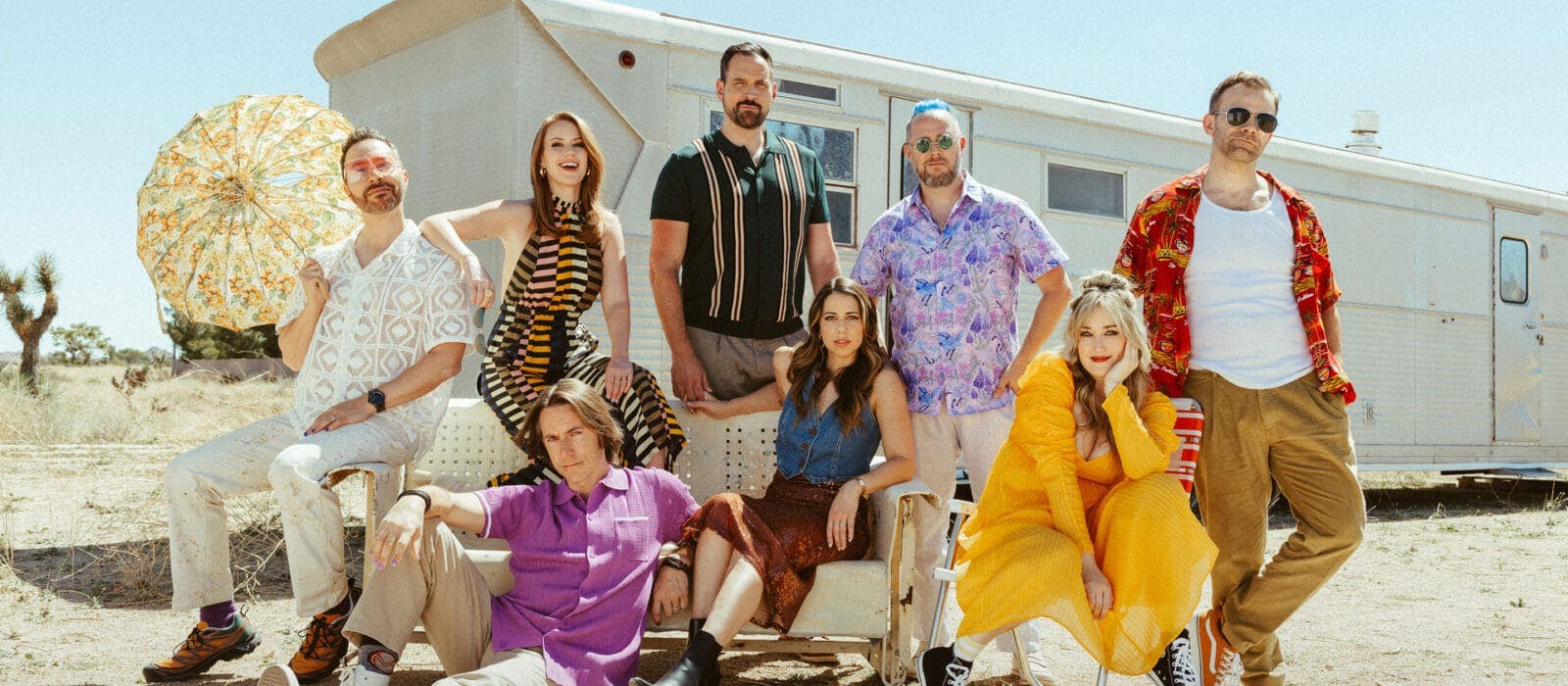 Ein Foto von Critical Role vor einem weißen Wohnwagen in einer Wüstenlandschaft: Von links nach rechts stehen und sitzen Sam mit einem gemusterten Sonnenschirm, Marisha in einem gestreiften Outfit, davor sitzt Matt in einem lila Hemd auf dem Boden, Travis in einem dunklen Poloshirt, Laura in einem Jeansoutfit, Taliesin in einem lilafarbenen Blumenhemd, Ashley in einem gelben Kleid und Liam in einem roten Hawaiihemd und Sonnenbrille. Alle tragen sommerliche, farbenfrohe Outfits.