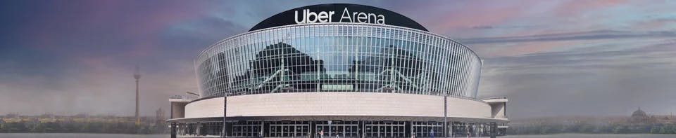 Die Uber Arena in Berlin: Ein modernes, rundes Veranstaltungsgebäude mit einer geschwungenen Glasfassade und dem Schriftzug „Uber Arena" auf dem Dach. Im Hintergrund ist die Berliner Stadtsilhouette unter einem lila-grauen Abendhimmel zu sehen.