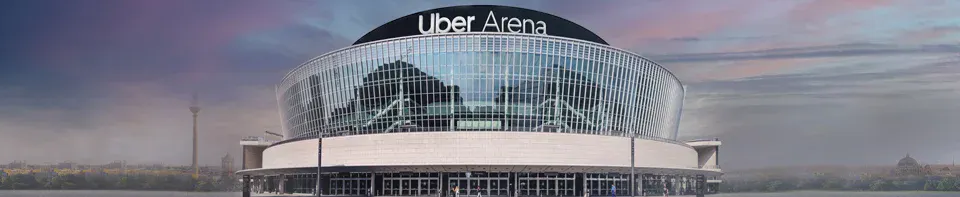Die Uber Arena in Berlin: Ein modernes, rundes Veranstaltungsgebäude mit einer geschwungenen Glasfassade und dem Schriftzug „Uber Arena" auf dem Dach. Im Hintergrund ist die Berliner Stadtsilhouette unter einem lila-grauen Abendhimmel zu sehen.