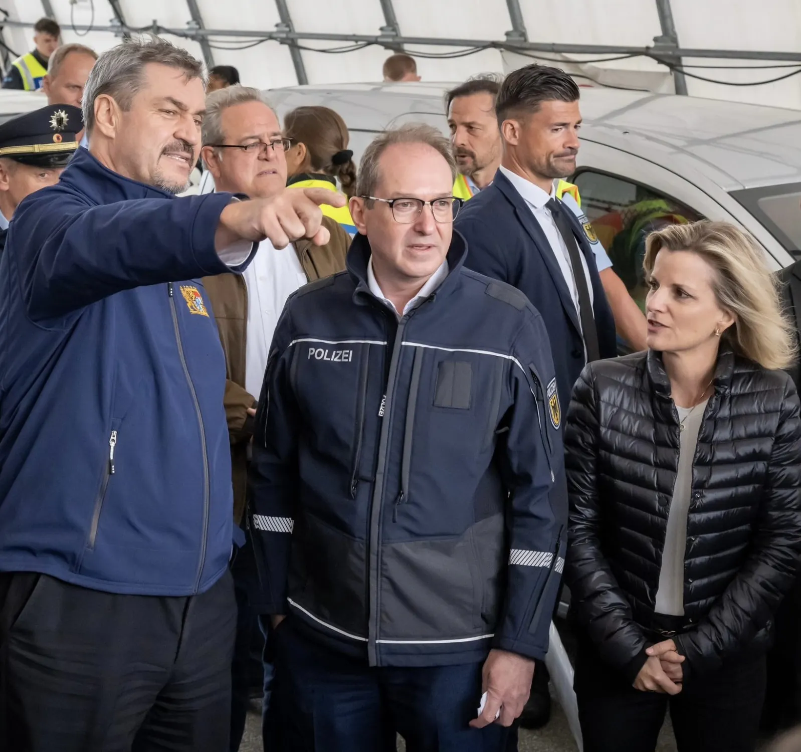 Markus Söder im Bayern-Fleece zeigt aus dem Bildrand heraus. Horst Dobrindt in Polizeijacke schaut unwirsch. Die ehemalige Broccolibeauftragte in Softshelljacke schaut interessiert auf Söders Pranke.
