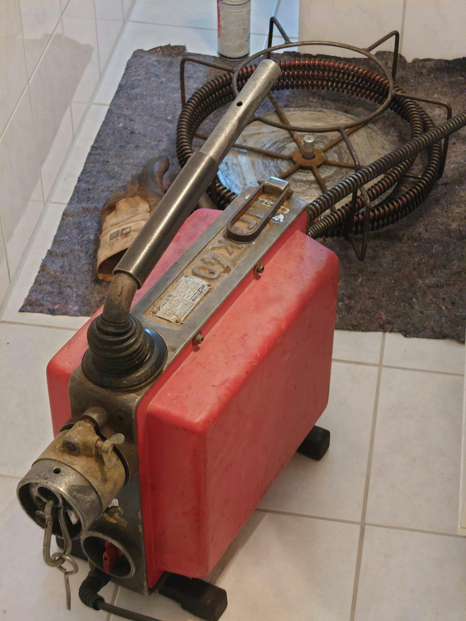 Ein Foto einer Rohrreinigungsmaschine. Ein Koffergroßer Klotz mit Plastikverkleidung und rundem Anschluss hinten. Vorne quillt eine lange Metallspirale heraus, etwa so dick wie Gartenschlauch. Obendrauf ein langer, in Richtung der Spirale gerichteter Hebel. Er glänzt in der vorderen Hälfte, weil der da so oft "angefasst" wurde.