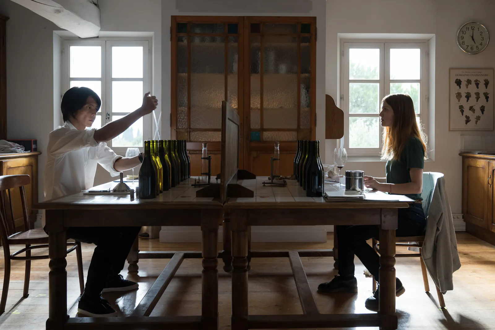 In dieser Filmszene von Drops of God sitzen sich die Schauspieler Tomohisa Yamashita und Fleur Geffrier an einem Tisch gegenüber, auf dem vor jedem hübsch aufgereiht Weinflaschen stehen.