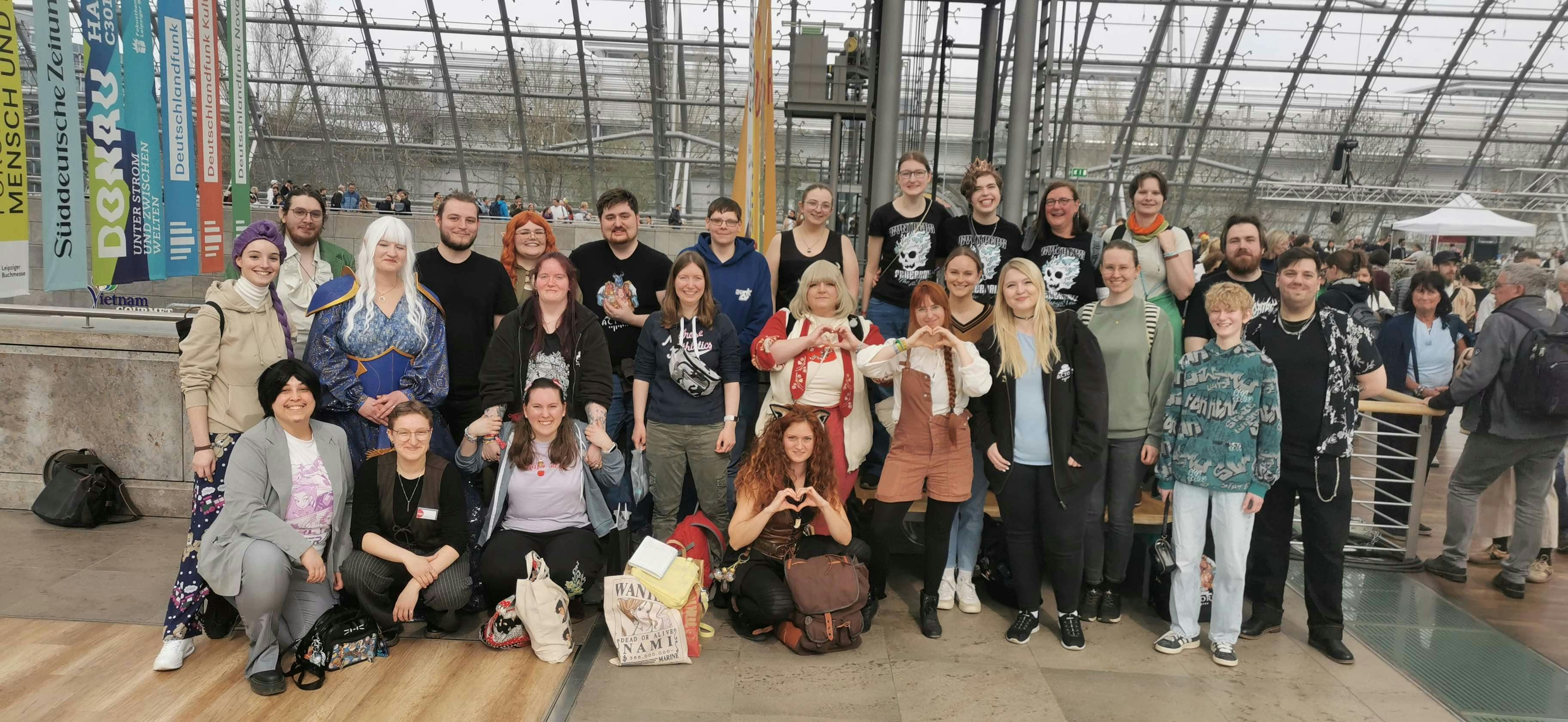 Ein Gruppenfoto von Diceys auf der Leipziger Buchmesse: Rund 25 Personen versammeln sich in einer großen Halle mit Glasdach und bunten Messefahnen im Hintergrund. Einige tragen Cosplays, andere Dice-Actors-Merchandise. Mehrere Personen formen Herzgesten mit ihren Händen. Die Gruppe strahlt gute Laune aus.