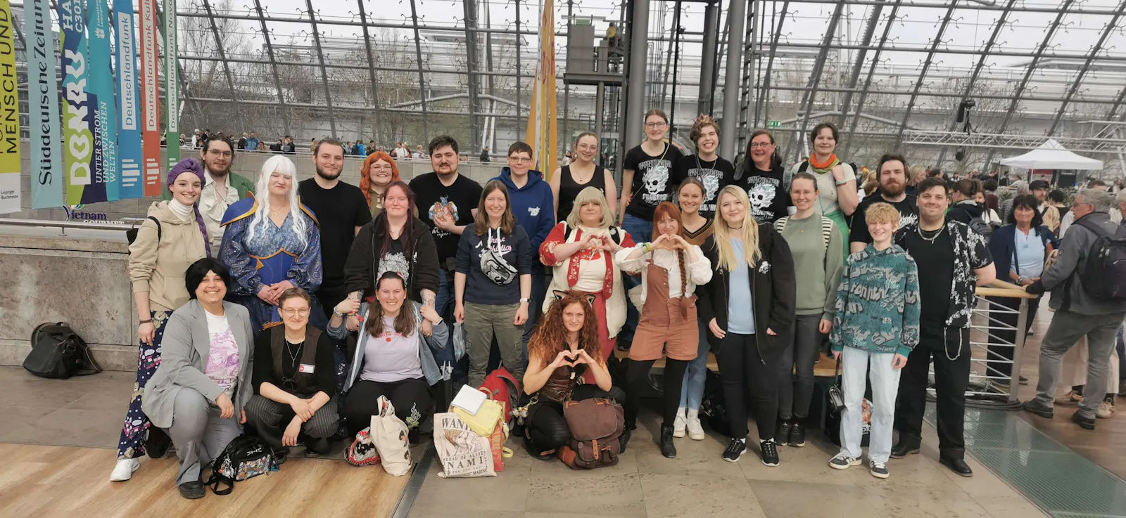 Ein Gruppenfoto von Diceys auf der Leipziger Buchmesse: Rund 25 Personen versammeln sich in einer großen Halle mit Glasdach und bunten Messefahnen im Hintergrund. Einige tragen Cosplays, andere Dice-Actors-Merchandise. Mehrere Personen formen Herzgesten mit ihren Händen. Die Gruppe strahlt gute Laune aus.