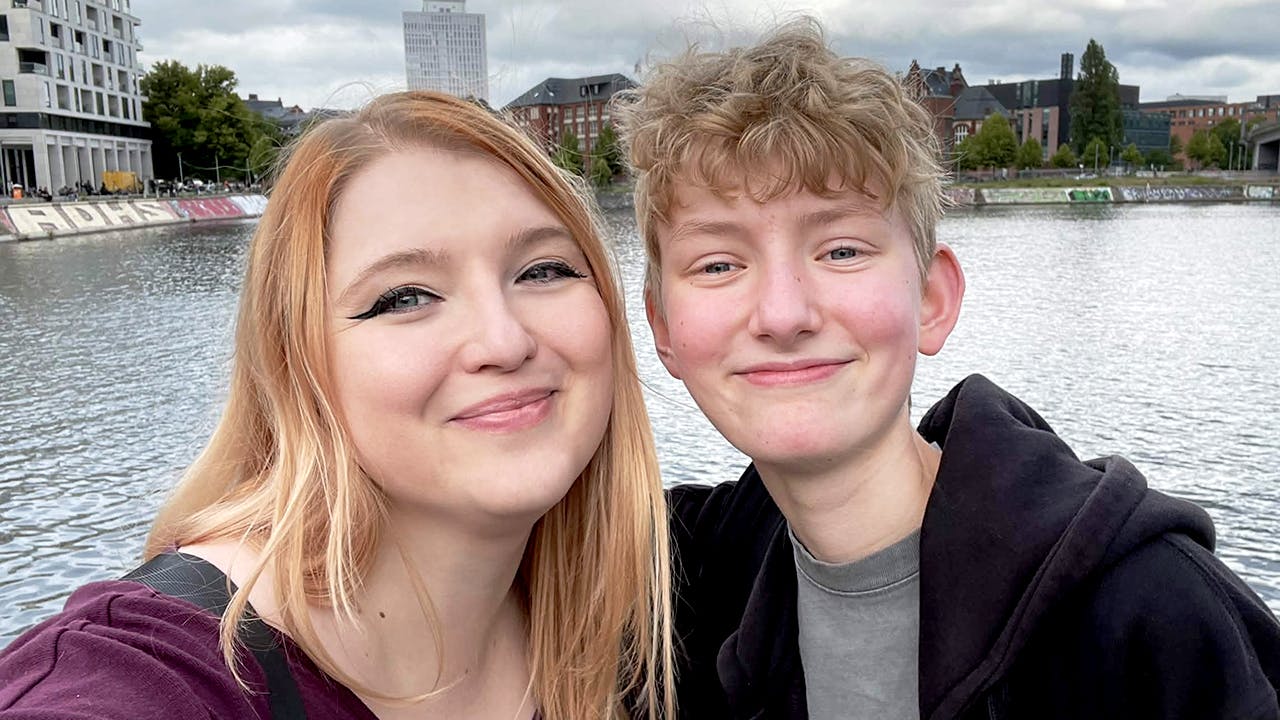 Ein Selfie von Rieke und Pauli am Humboldthafen in Berlin, direkt neben dem Hauptbahnhof. Links lächelt Rieke mit langen rotblonden Haaren und einem lila Oberteil in die Kamera. Rechts steht Pauli mit kurzem blondem Haar und einem schwarzen Dice-Actors-Hoodie und lächelt ebenfalls. Im Hintergrund sind das Hafenbecken und die Berliner Stadtsilhouette unter bewölktem Himmel zu sehen.