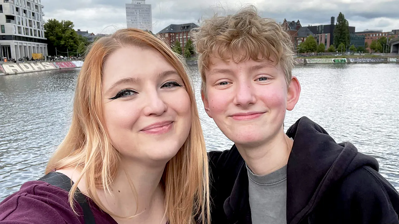 Ein Selfie von Rieke und Pauli am Humboldthafen in Berlin, direkt neben dem Hauptbahnhof. Links lächelt Rieke mit langen rotblonden Haaren und einem lila Oberteil in die Kamera. Rechts steht Pauli mit kurzem blondem Haar und einem schwarzen Dice-Actors-Hoodie und lächelt ebenfalls. Im Hintergrund sind das Hafenbecken und die Berliner Stadtsilhouette unter bewölktem Himmel zu sehen.