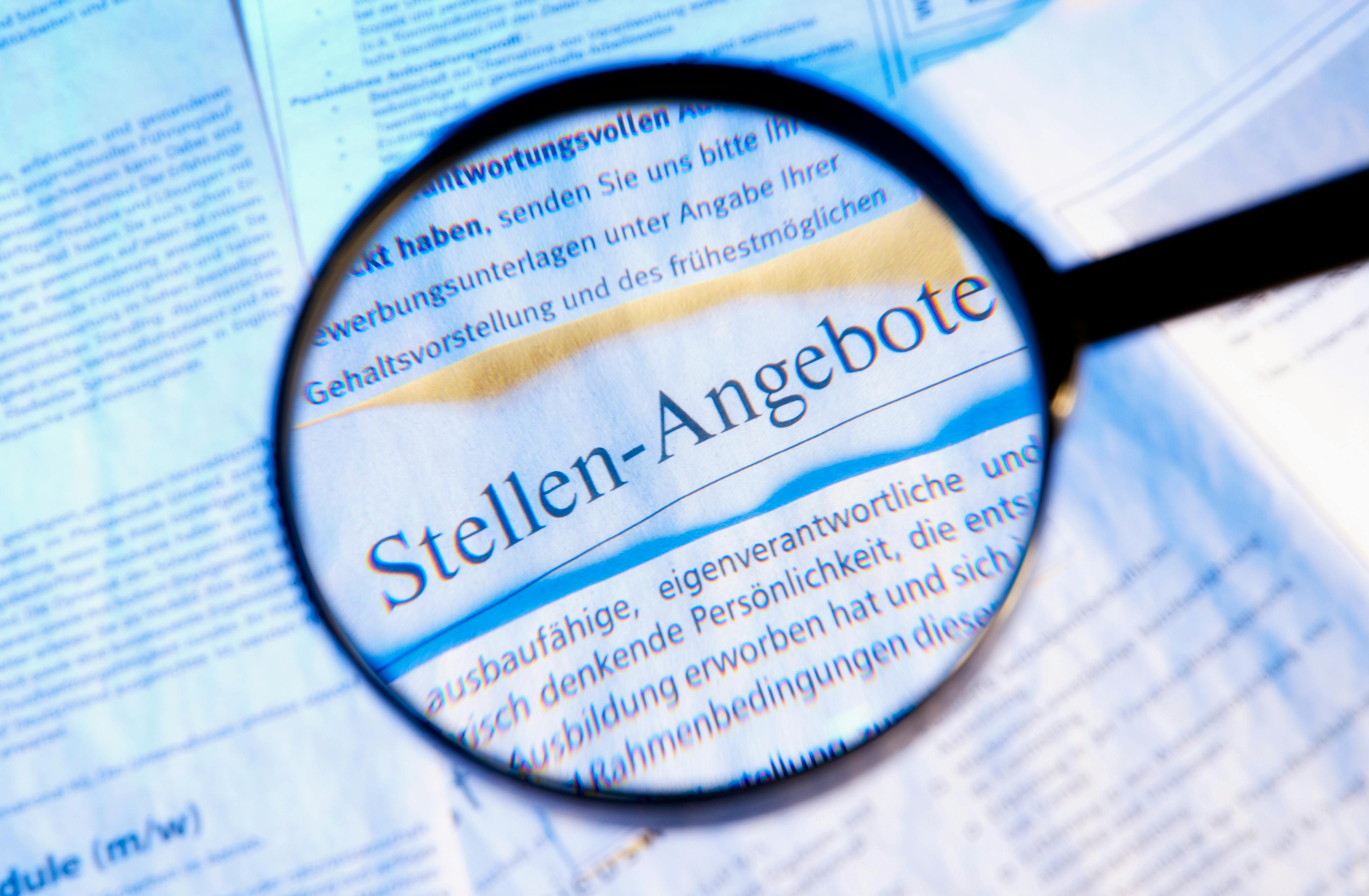 Das Foto zeigt eine Zeitungsseite mit Stellenanzeigen, die durch eine Lupe vergrößert wird, um die Details besser erkennen zu können.