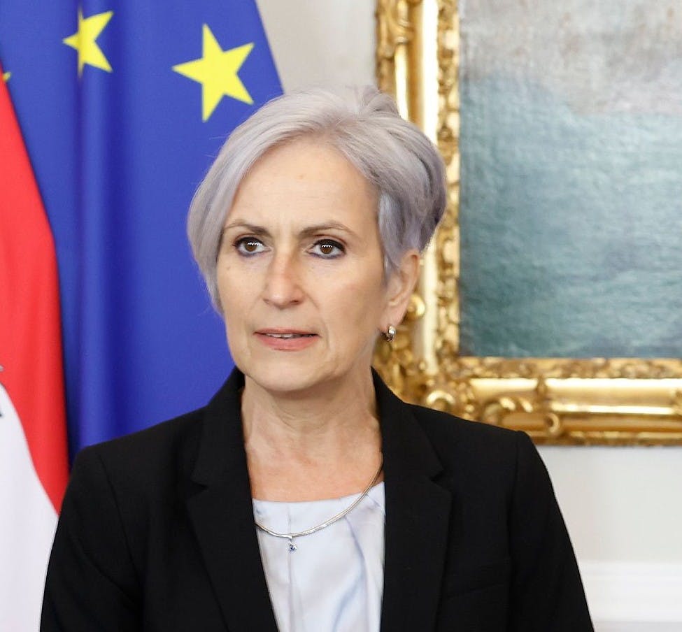 Auf dem Foto ist die Justizministerin Anna Sporrer zu sehen. Sie steht vor einer EU-Flagge und dem Rahmen eines Gemäldes.