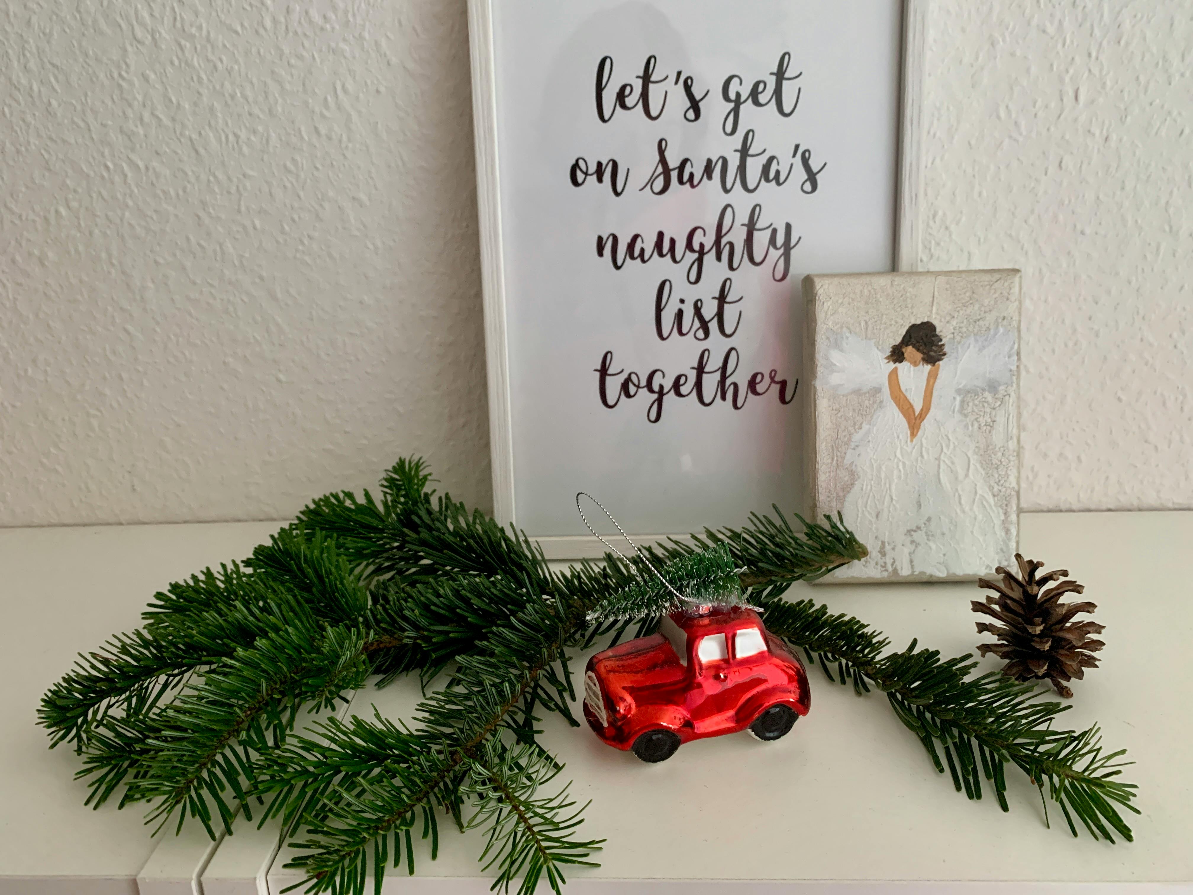 Auf einem weißen Sideboard liegt ein Tannenzweig, dazwischen steht ein rotes Auto als Christbaumschmuck. Dahinter stehen ein Bild mit der Aufschrift "Let's get on Santas naughty List together" und ein Bild von einem Engel.