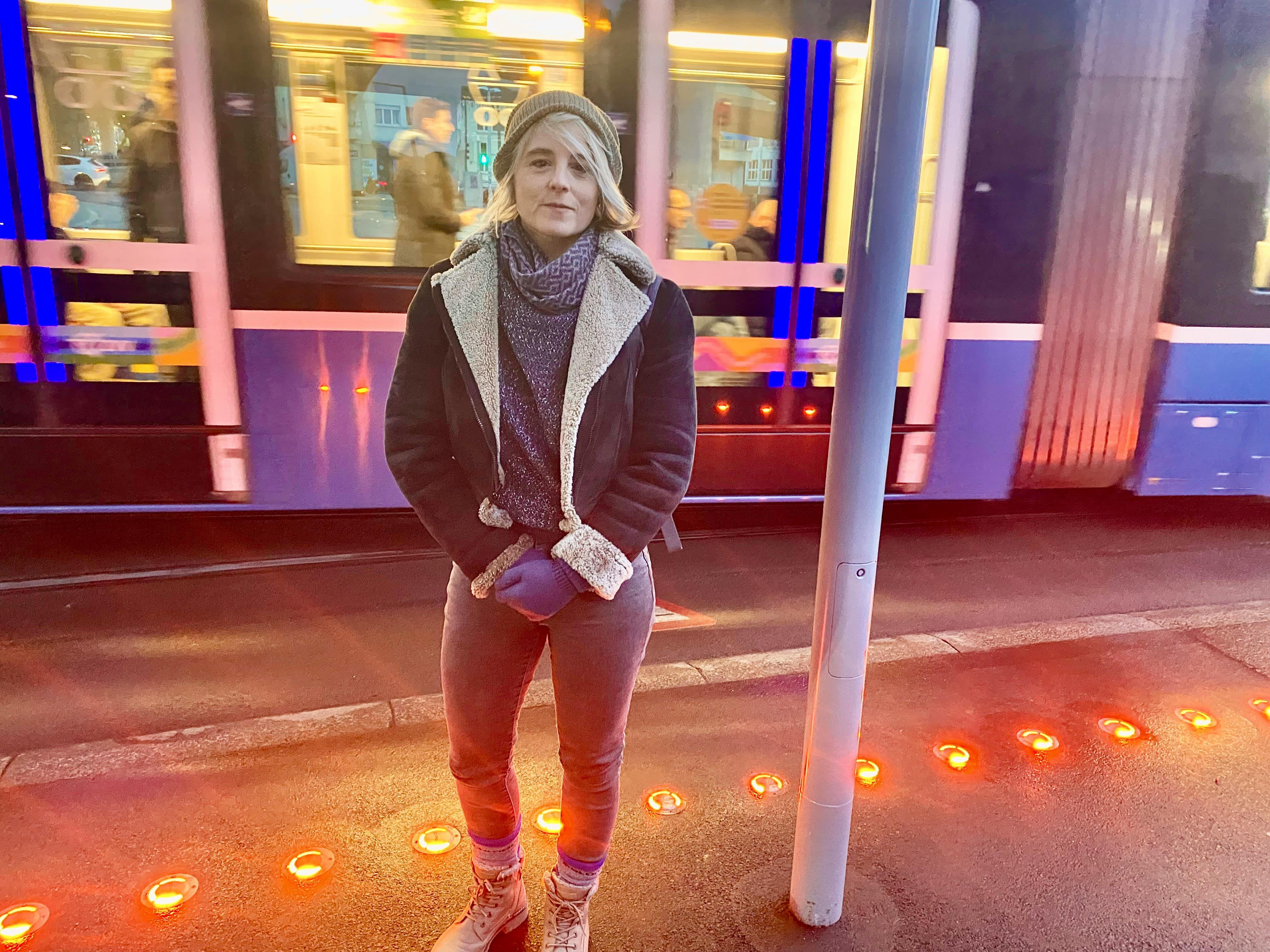 Das Bild zeigt die Politikerin Sandra Bienek auf dem Escher-Wyss-Platz in Zürich. Im Hintergrund ein durchfahrendes Tram, vorne am Boden rote Warnlichter.