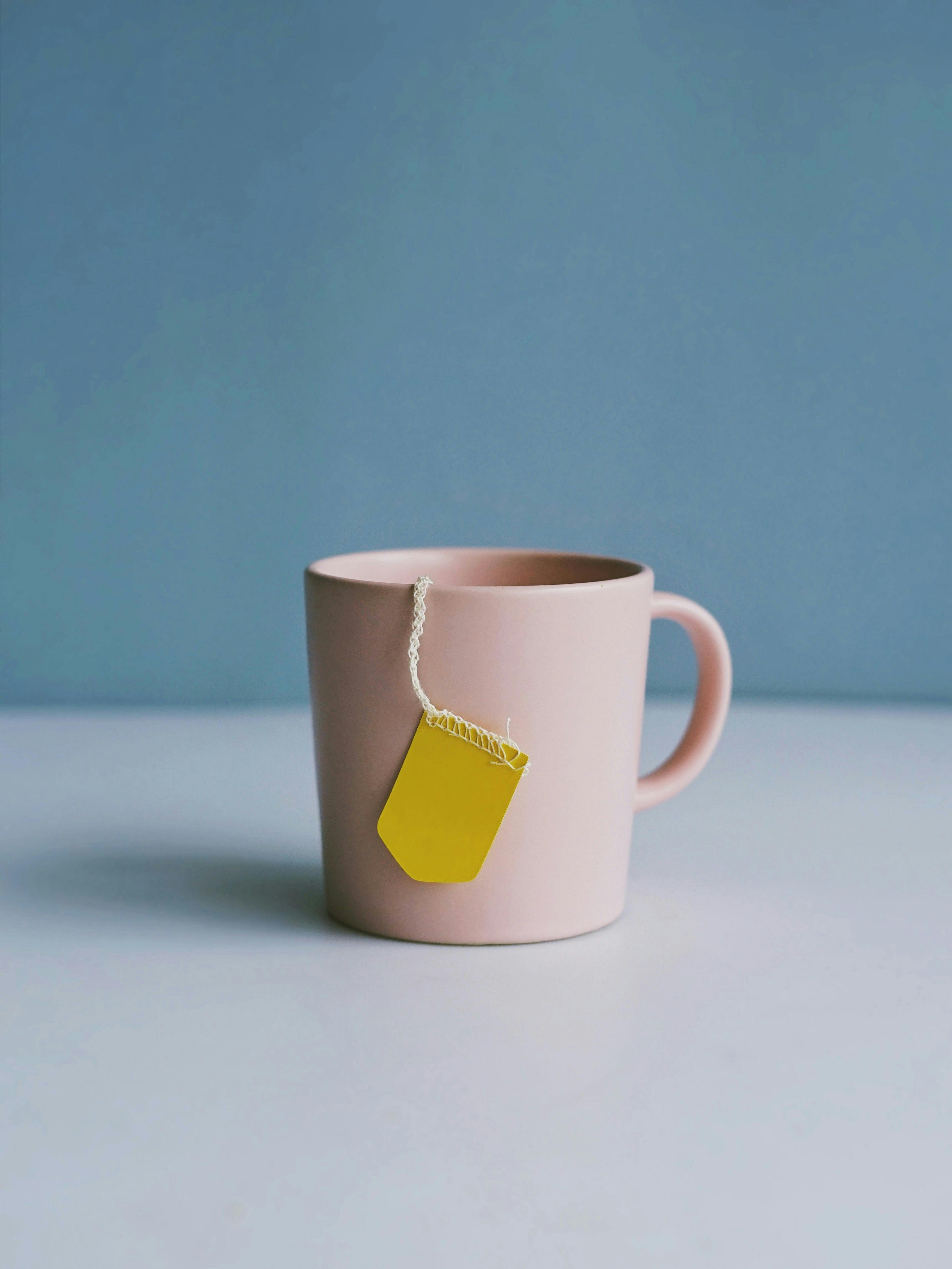 Eine rosafarbene Tasse vor blauem Hintergrund. Ein Bändchen von einem Teebeutel mit gelbem Etikett hängt über den Rand.