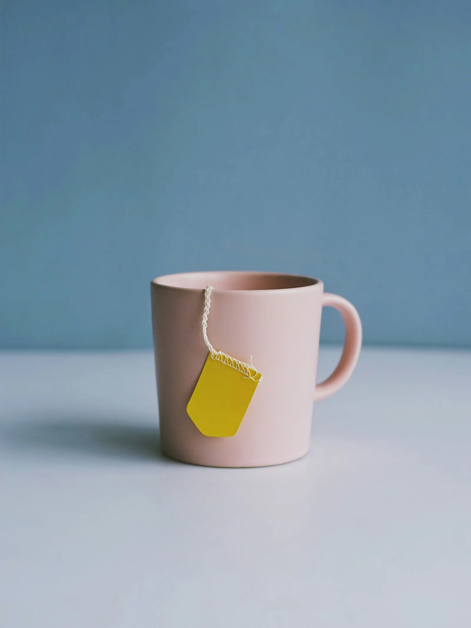 Eine rosafarbene Tasse vor blauem Hintergrund. Ein Bändchen von einem Teebeutel mit gelbem Etikett hängt über den Rand.
