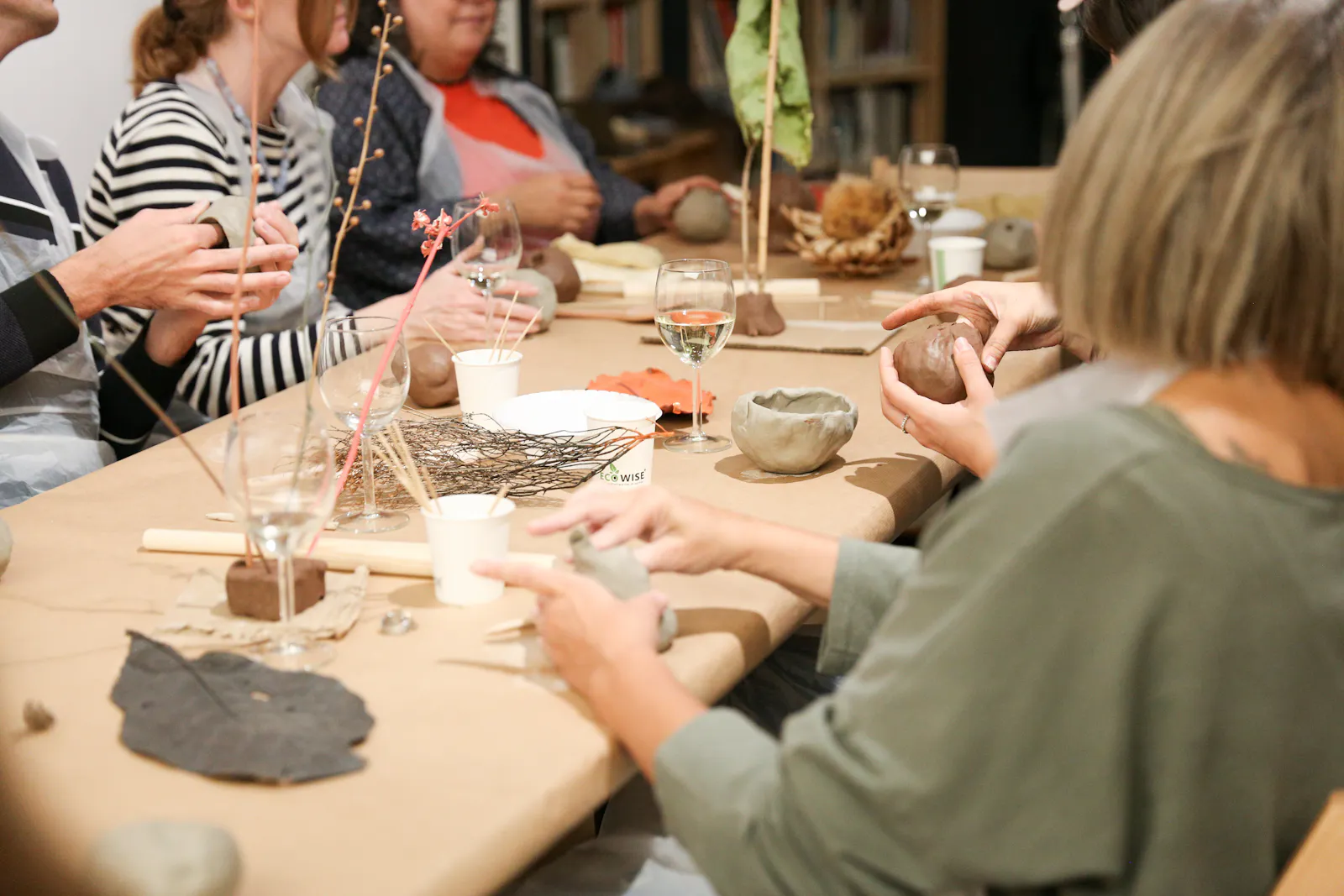 Image of people in a clay-making workshop. Five people are sat around a table wearing white plastic aprons and moulding pieces of clay in their hands. On the table, there are dried flowers, plants and leaves.