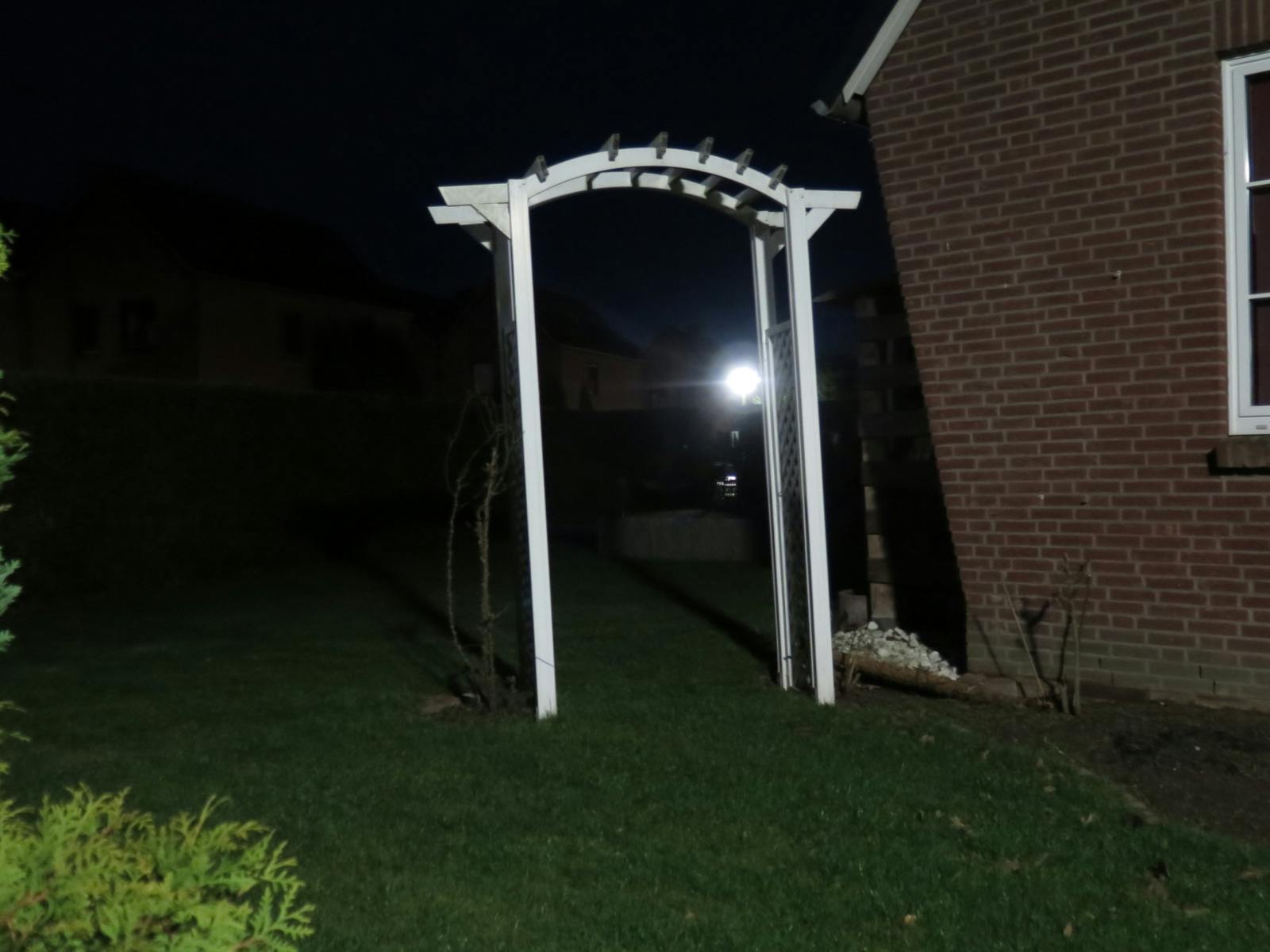 Nacht, Rasen, darauf ein seltsamer weißer Torbogen. Rechts davon angeschnitten die Rotklinkermauer eines Hauses.