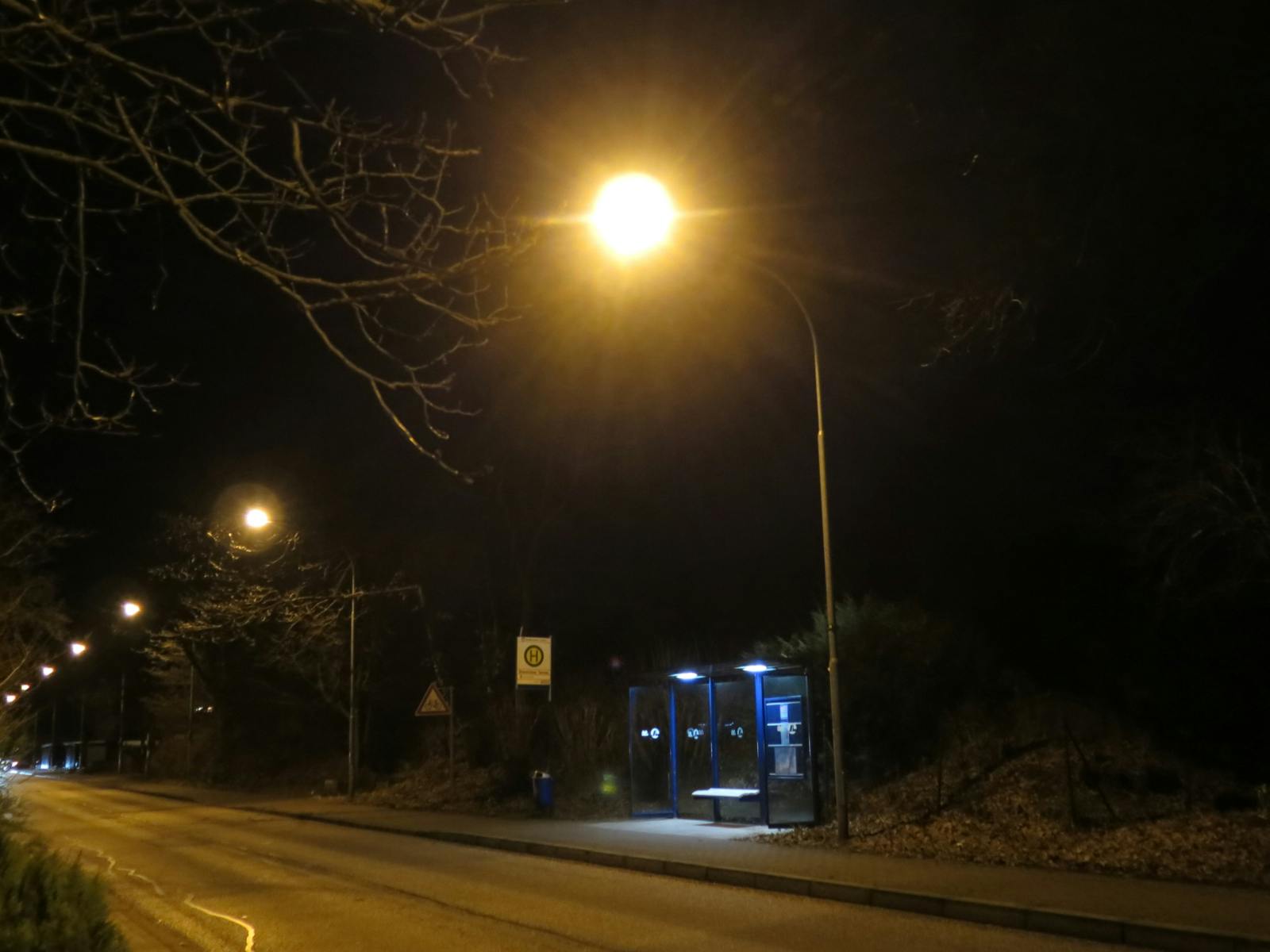 Eine verlassene Straße in der Nacht, gelbe Straßenlaternen, an einer Böschung eine einsame Bushaltestelle.