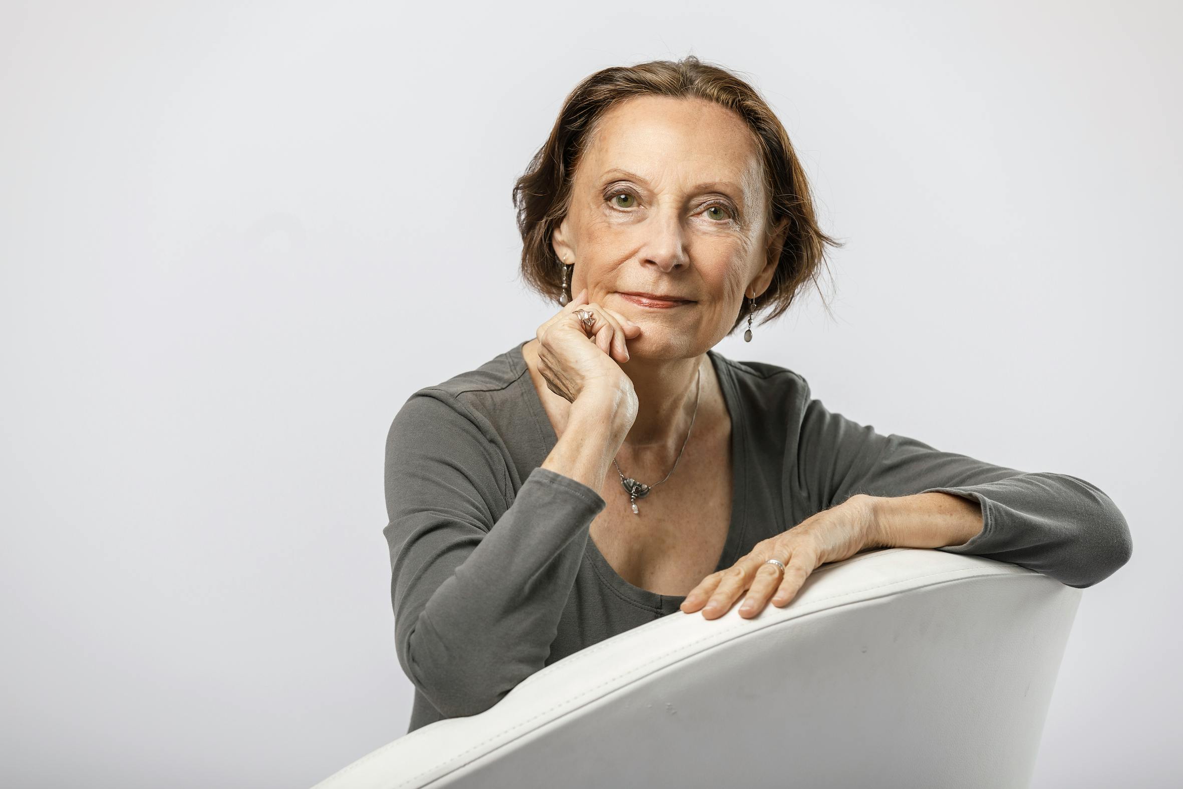 Eine mittelalte Frau mit braunen, halblangen Haaren und einem grauen Oberteil sitz auf einem weißen Sessel.