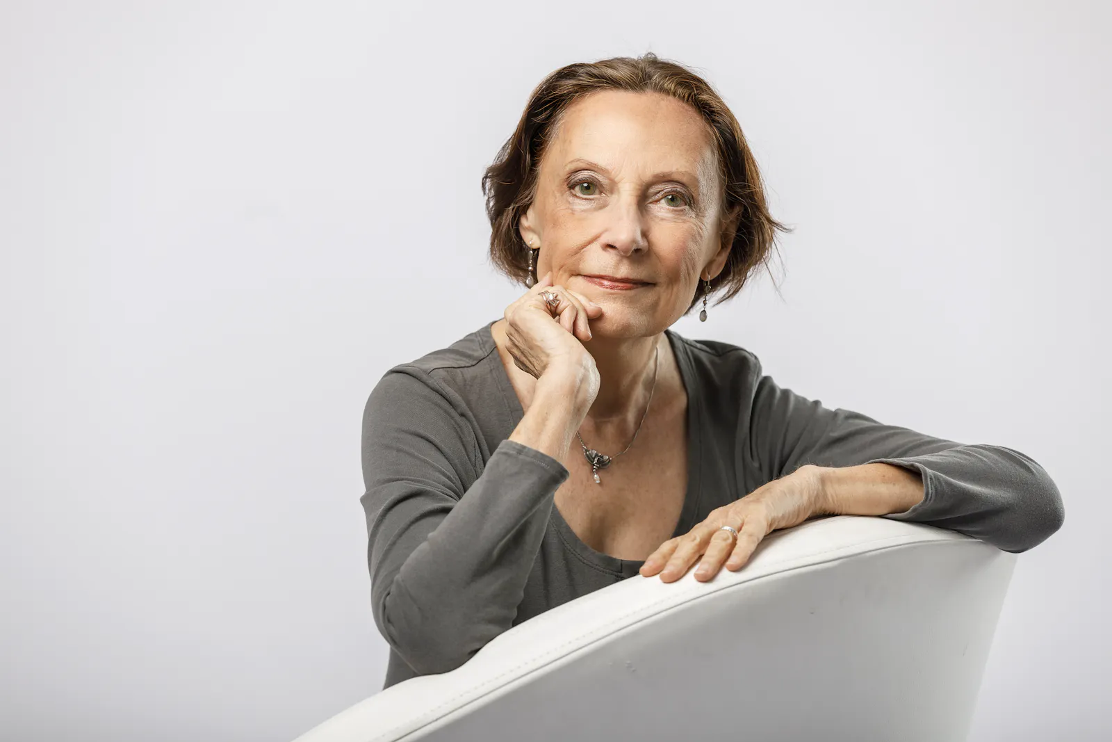 Eine mittelalte Frau mit braunen, halblangen Haaren und einem grauen Oberteil sitz auf einem weißen Sessel.