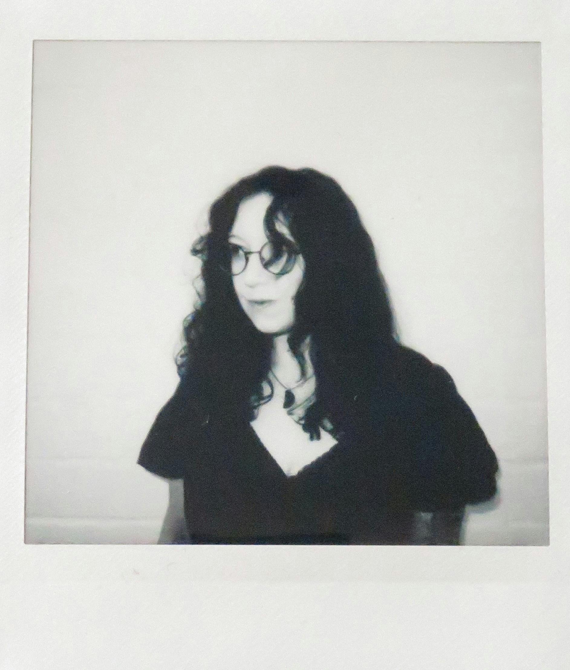 Schwarz weiß Fotografie einer Frau mit langen dunklen Haaren. Sie trägt eine Brille und blickt links aus dem Bild heraus. Der Hintergrund ist eine weiße Wand.